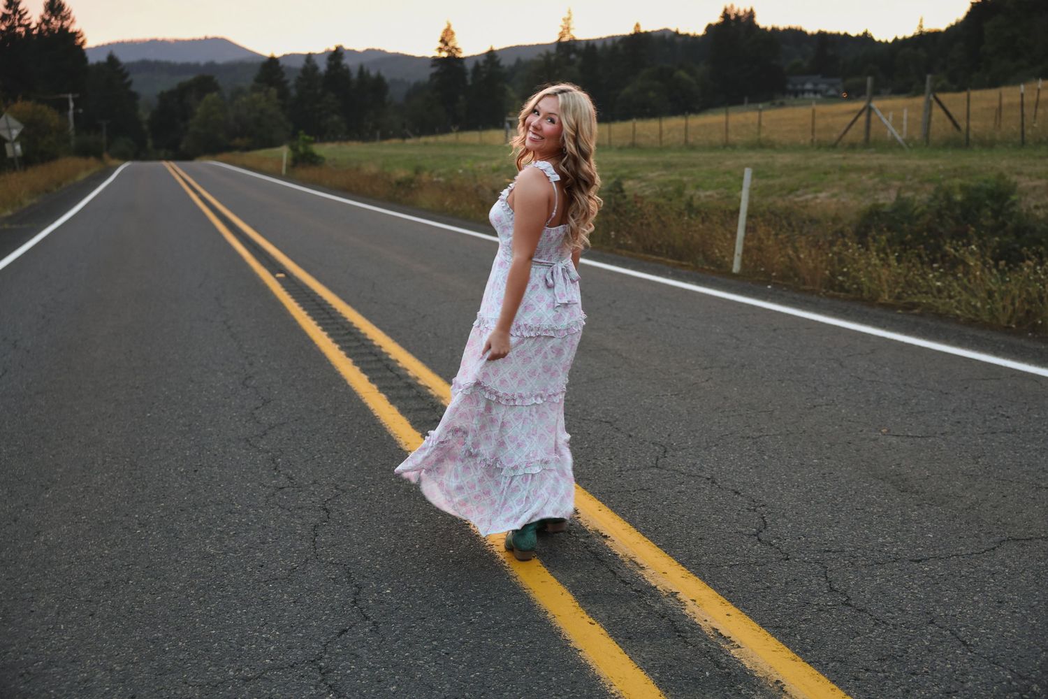 Blonde woman in floral maxi dress standing on rural road with mountain scenery at dusk.