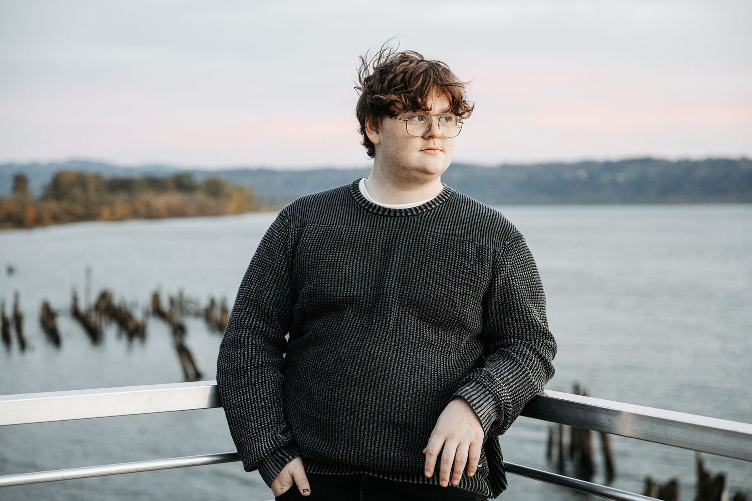 Young person in gray knit sweater poses on waterfront deck with misty river and wooden pilings behind.