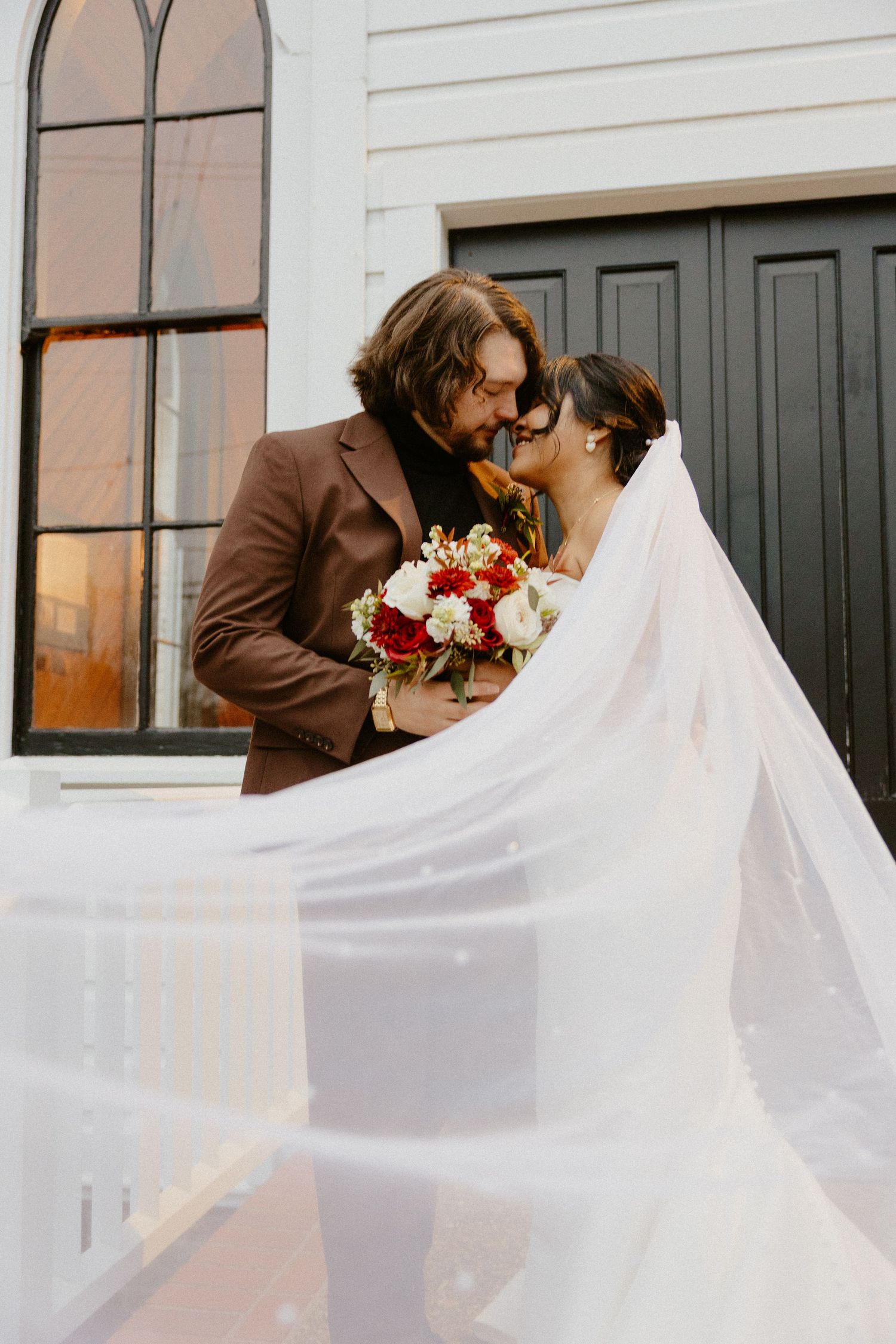 Bride and groom sharing a romantic moment, her veil flowing, holding red and white bouquet before a white chapel.