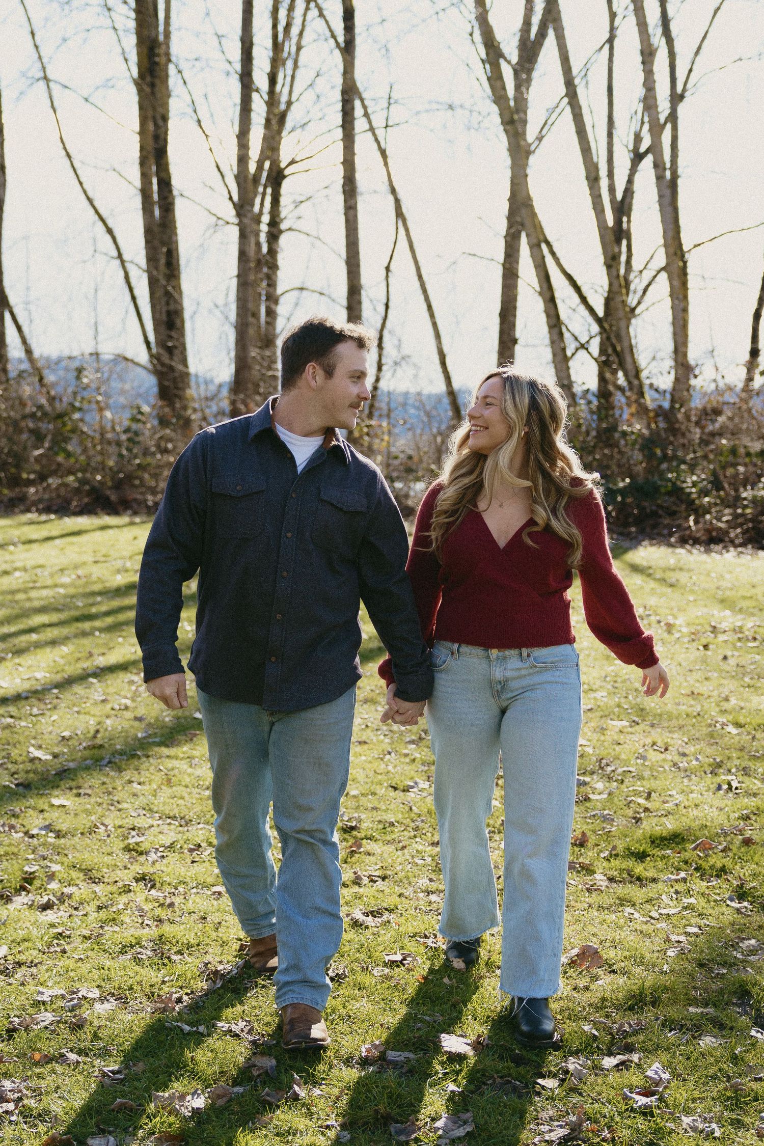 Smiling couple holding hands and walking together on green grass with bare winter trees.