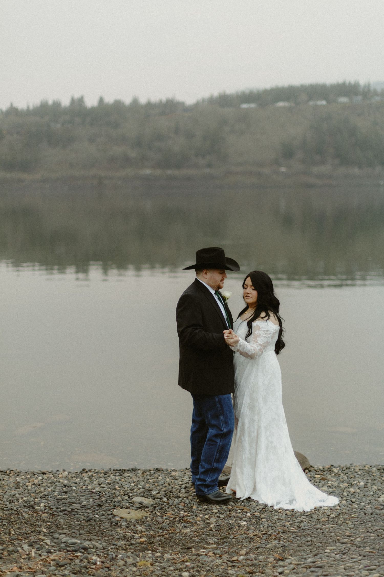 Bride and groom sharing a romantic lakeside moment, groom in black cowboy hat and suit, misty hills behind.