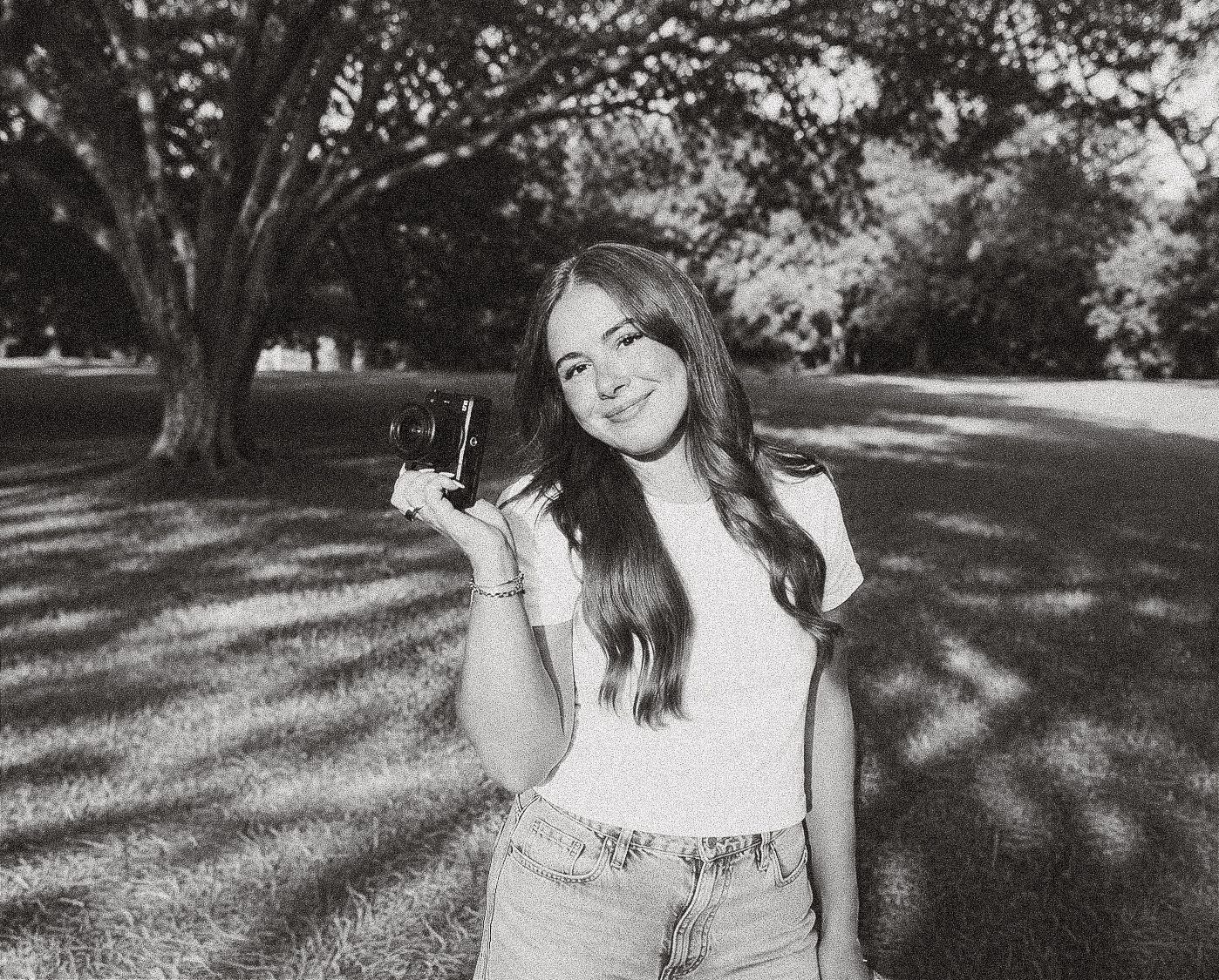 Young woman smiling in a park holding a camera, black and white portrait photo outdoors.
