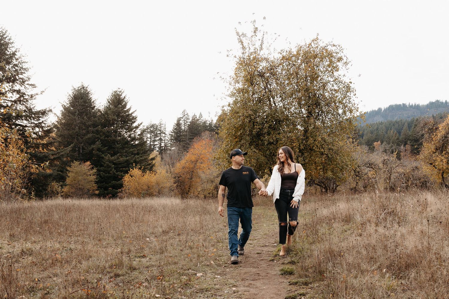Couple holding hands walking on a dirt path through autumn meadow with colorful fall trees.