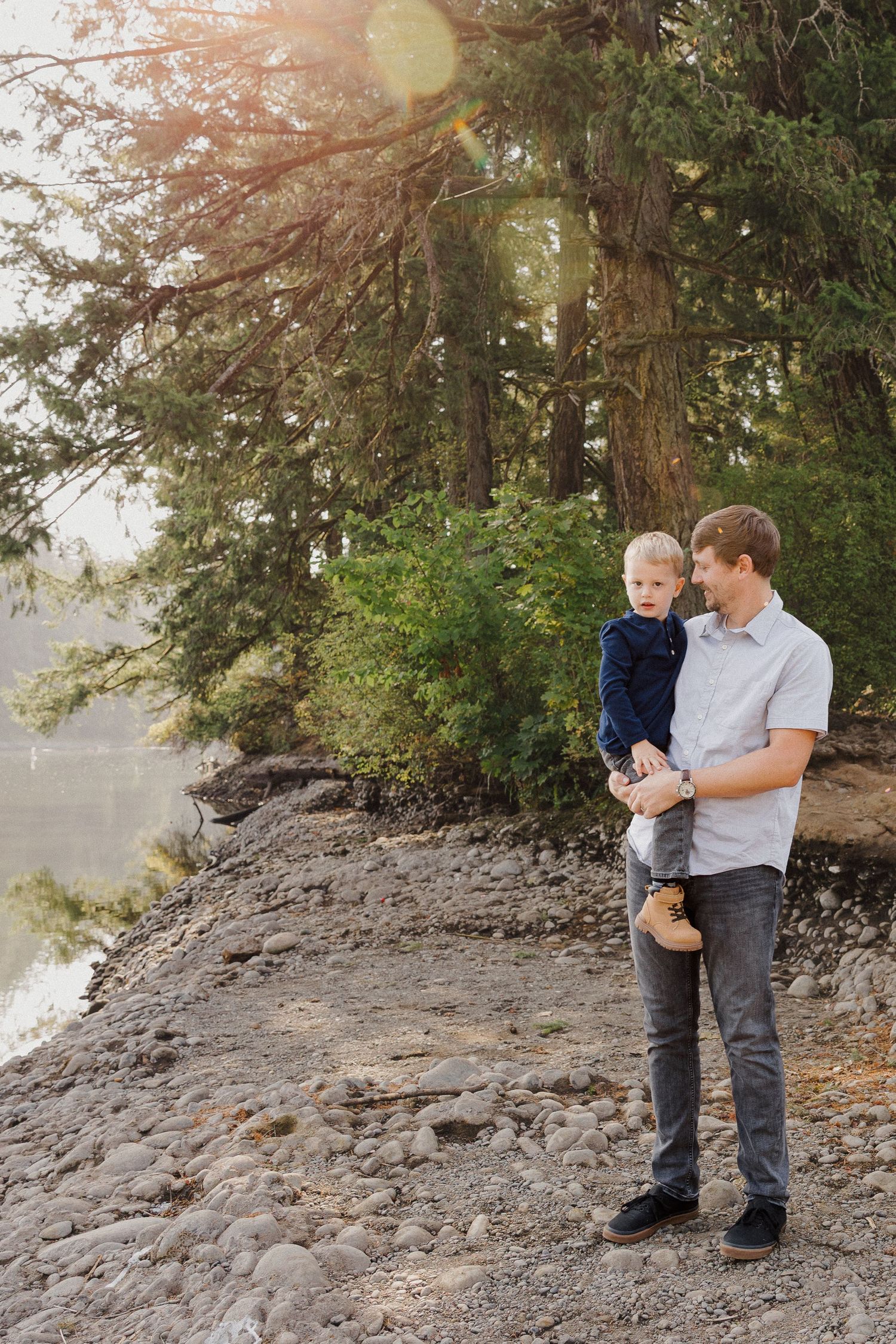 Father holding young toddler son on rocky lakeside shore surrounded by pine trees.