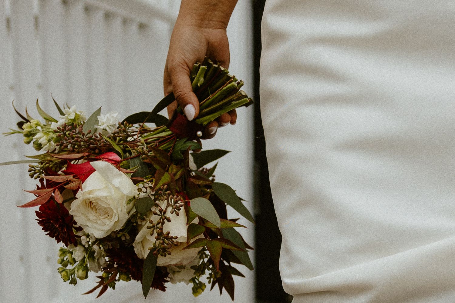 Bride in white dress holding a lush bouquet of burgundy, white, and green flowers with dark ribbon.