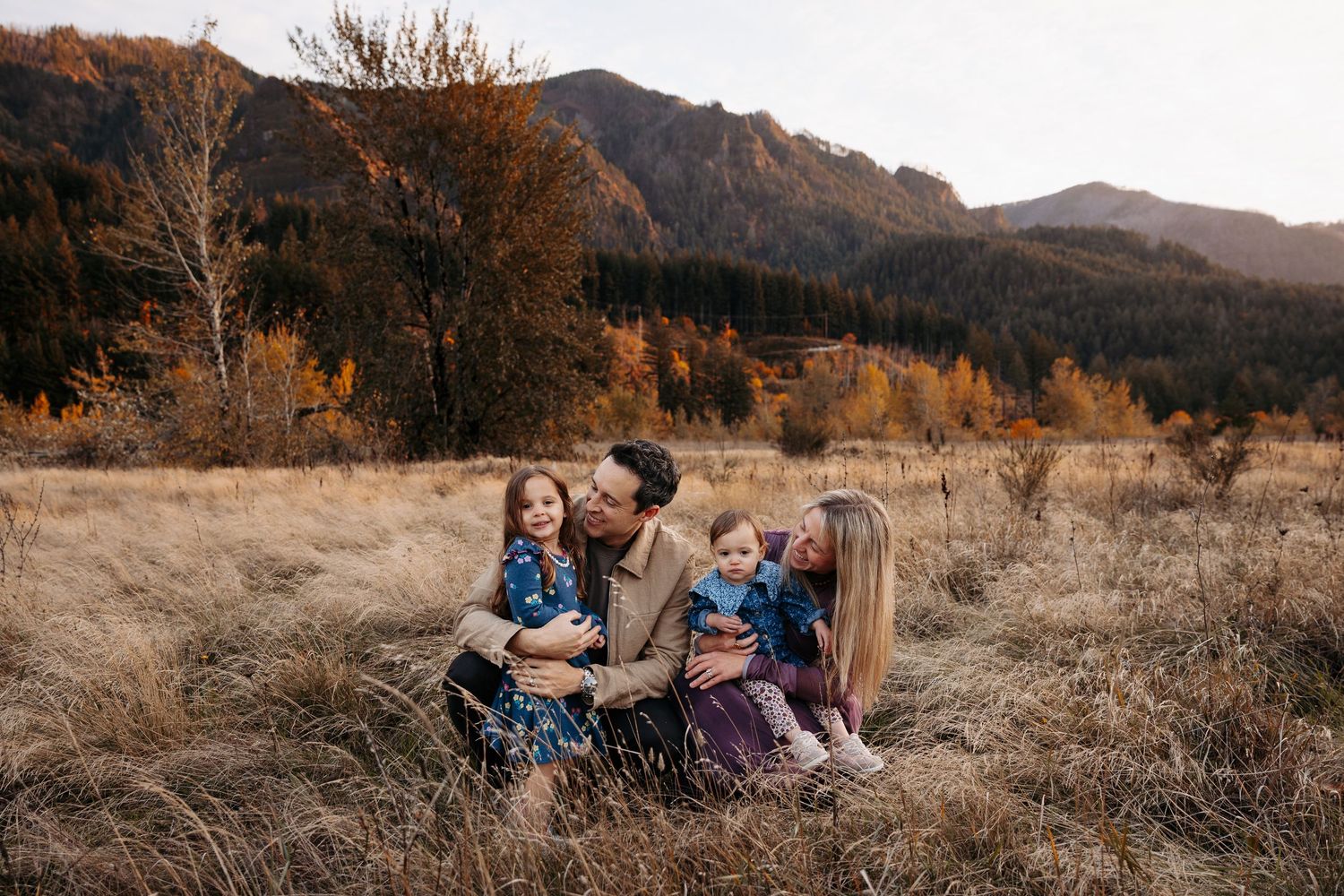 A happy family of four sits together in a golden autumn meadow with mountain backdrop.
