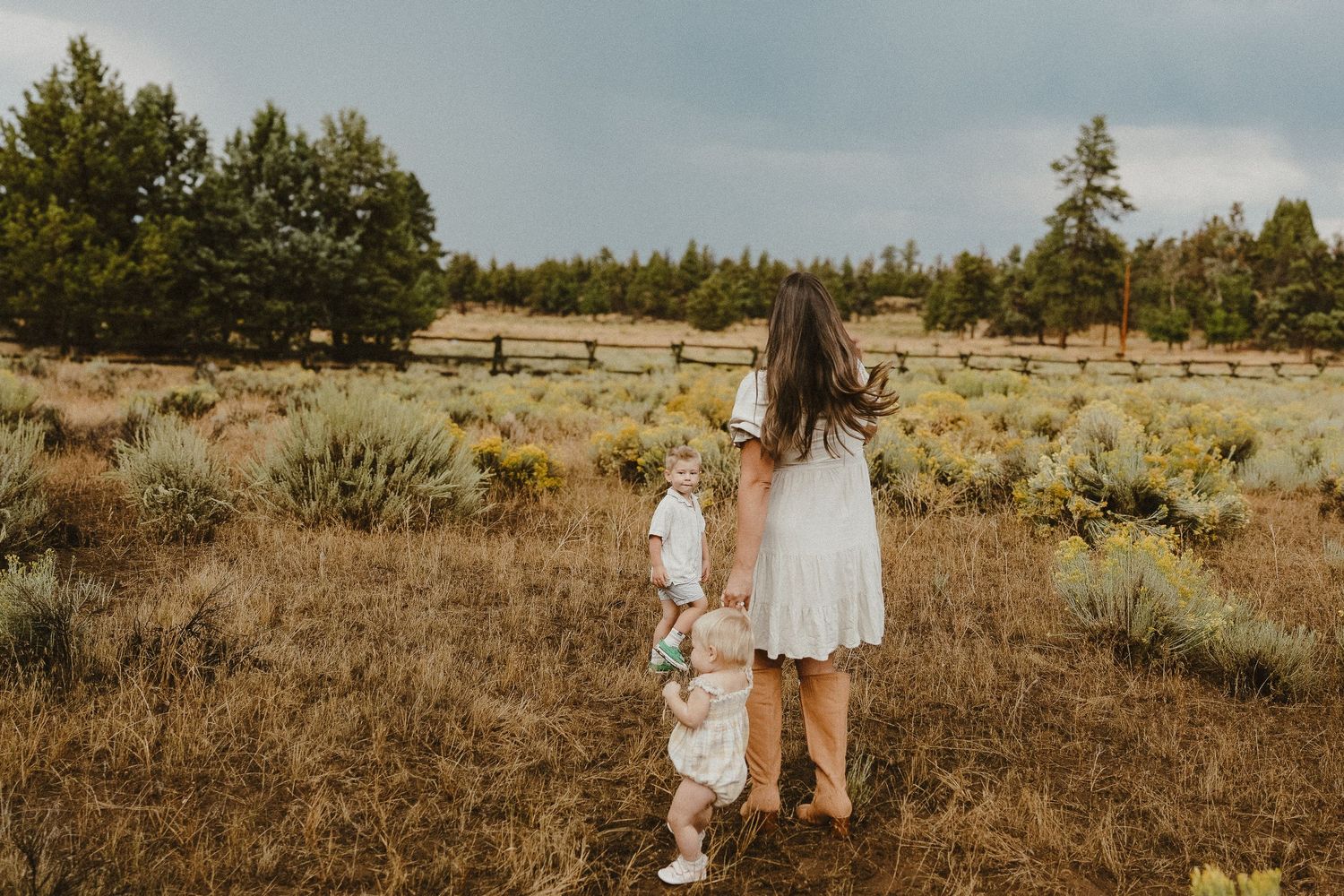 Mother in white dress walks with two young children through a scenic meadow with pine trees.