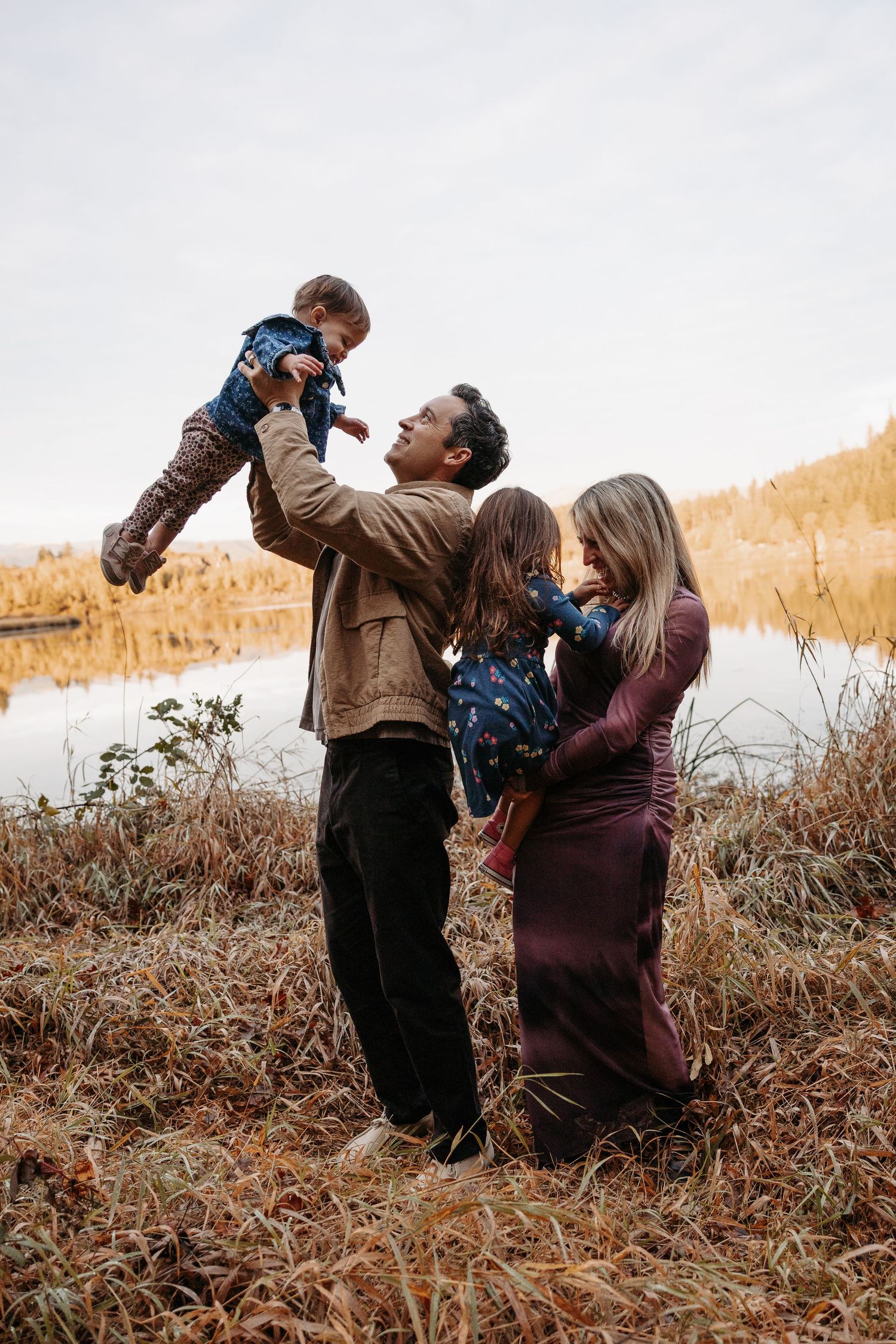 Father lifting baby joyfully while mother and daughter embrace near autumn lake in golden field.
