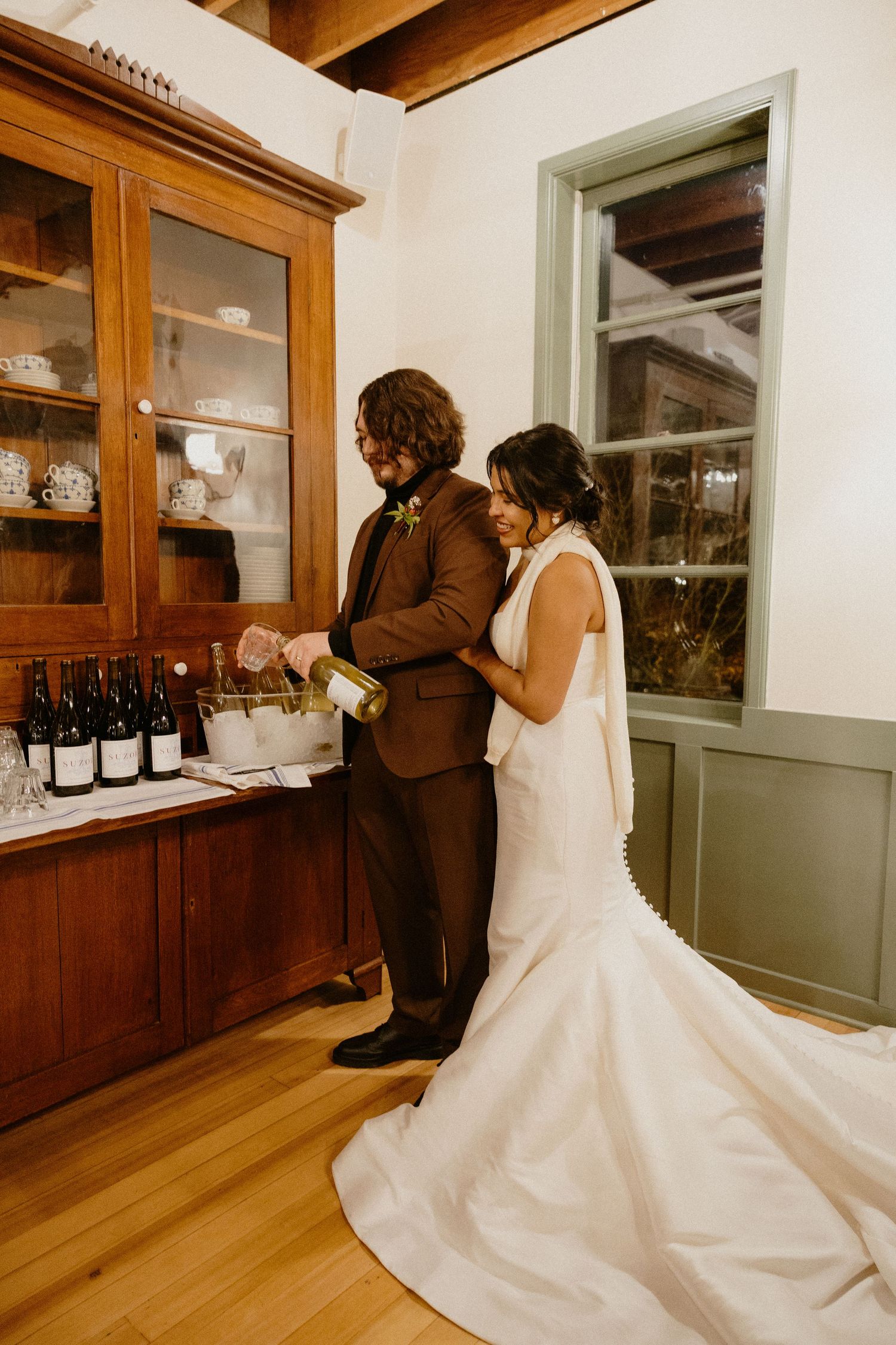 Bride and groom pouring wine together at wooden cabinet during wedding reception.