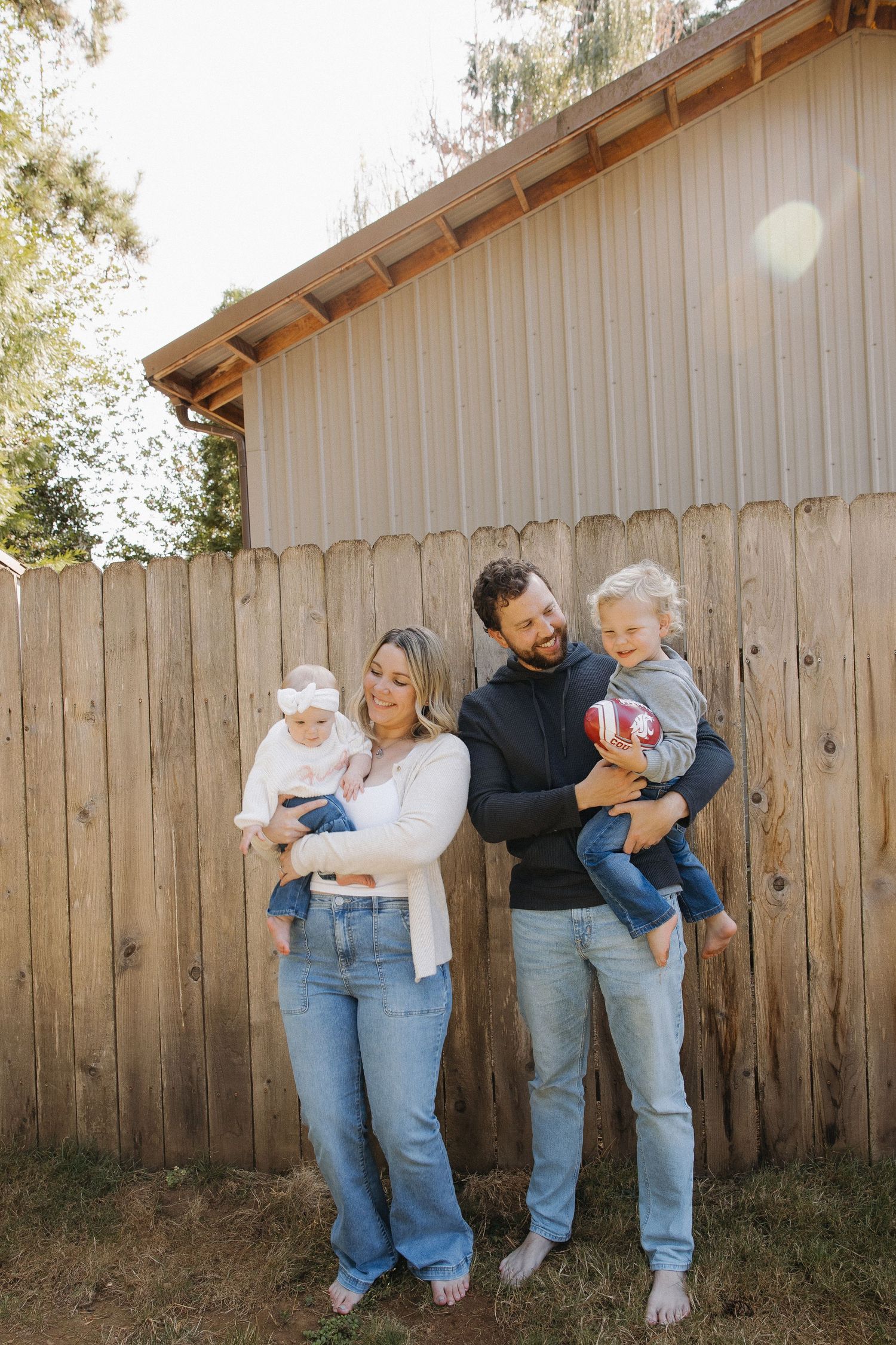 Family of four standing outdoors holding two young children against wooden fence backdrop.