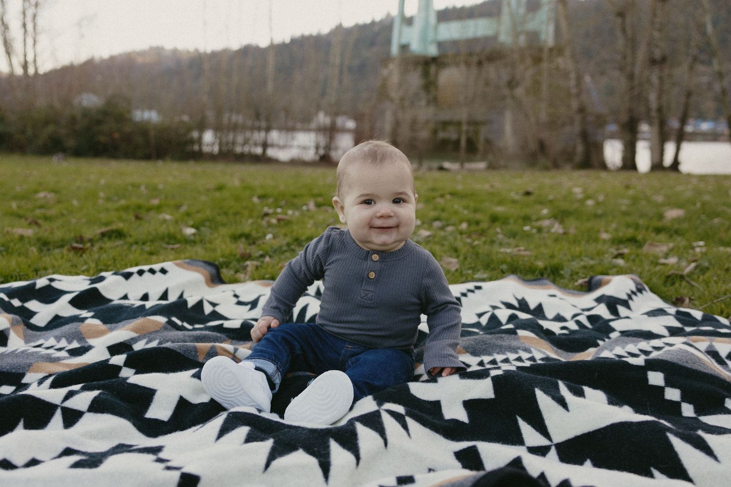 Happy baby boy sitting on geometric patterned blanket on grass with St. Johns Bridge in background.