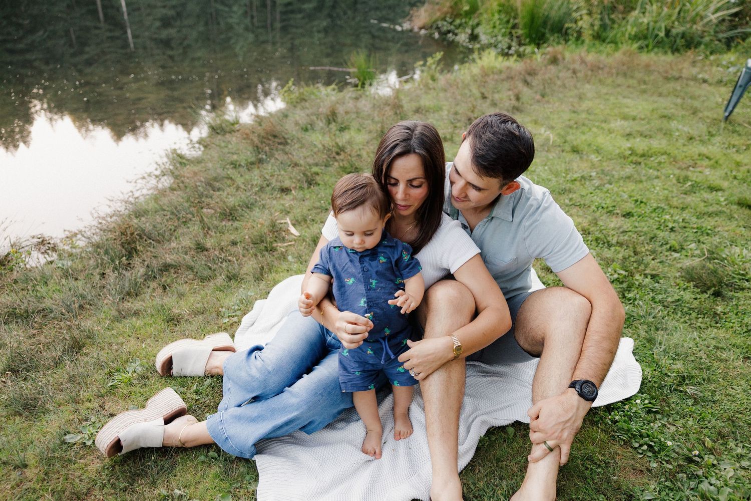Happy family of three sitting on a blanket by a peaceful pond in a lush forest.