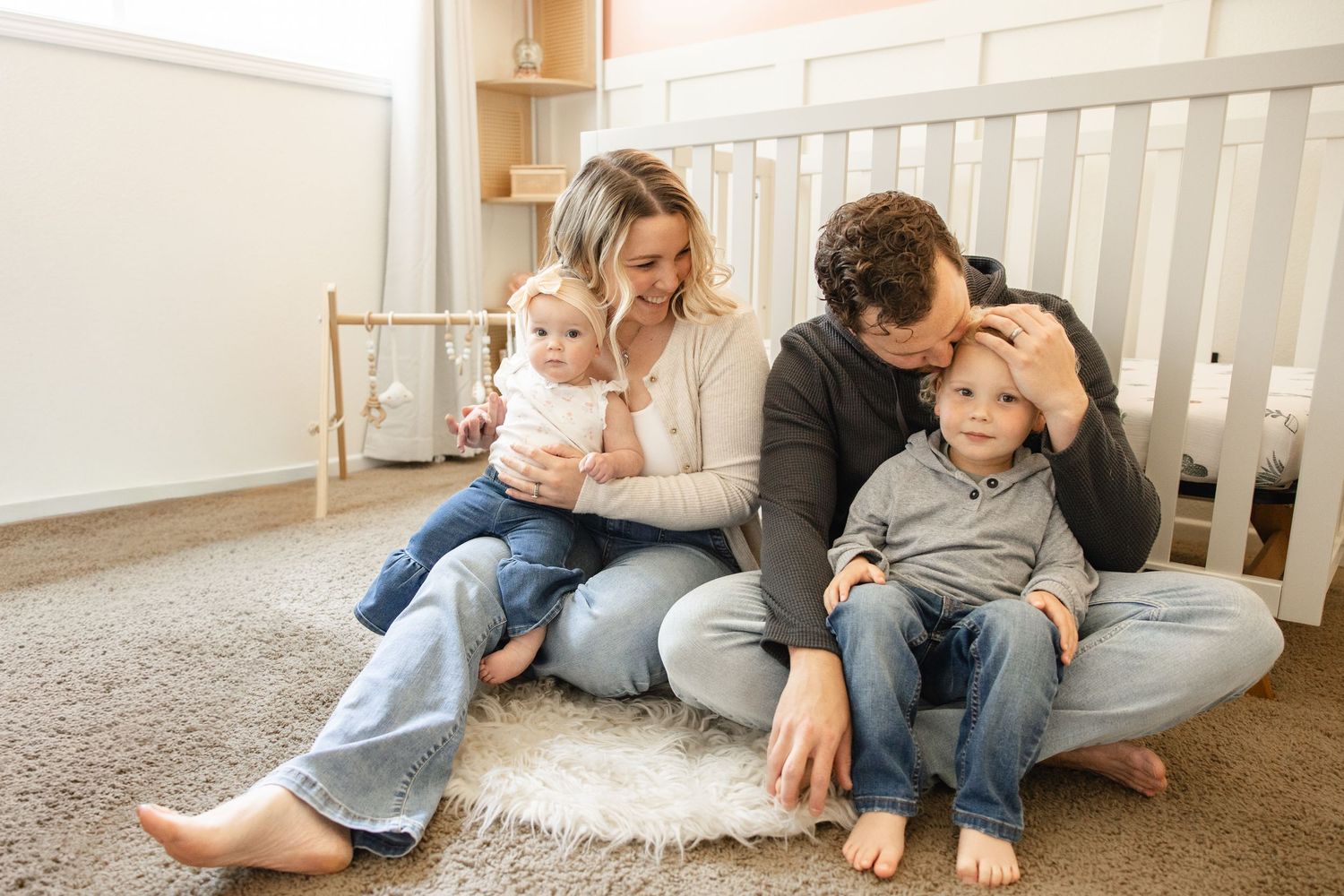 Family of four sitting on nursery floor, parents tending to baby and toddler near white crib.
