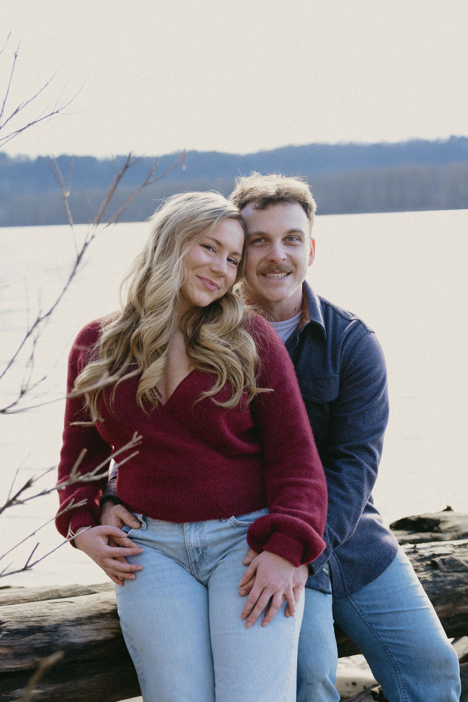 Couple posing by a lake, woman in red sweater and jeans leaning against man in navy shirt sitting on a log.