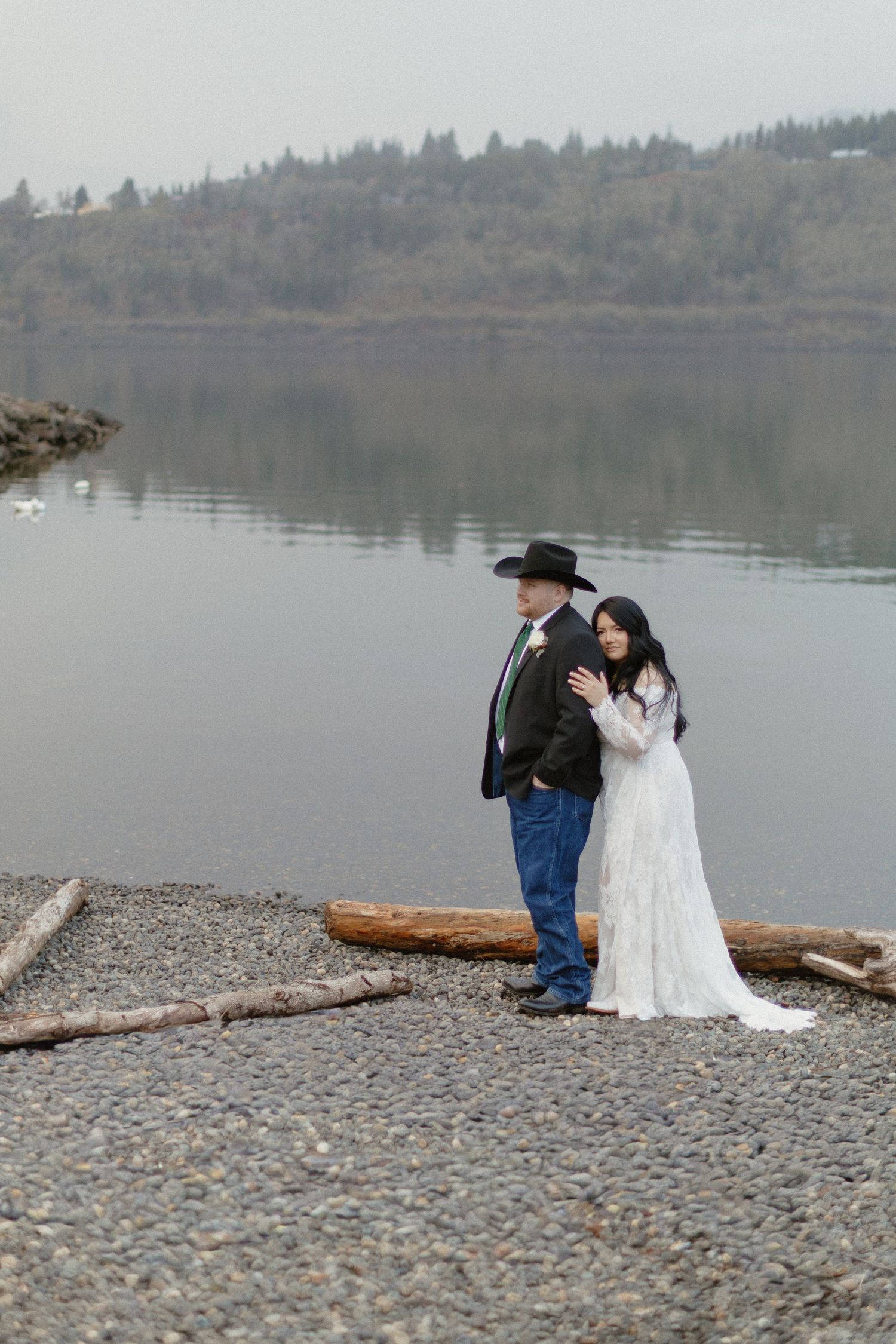 Wedding couple embracing on a misty lakeside beach with driftwood and forested hills behind them.