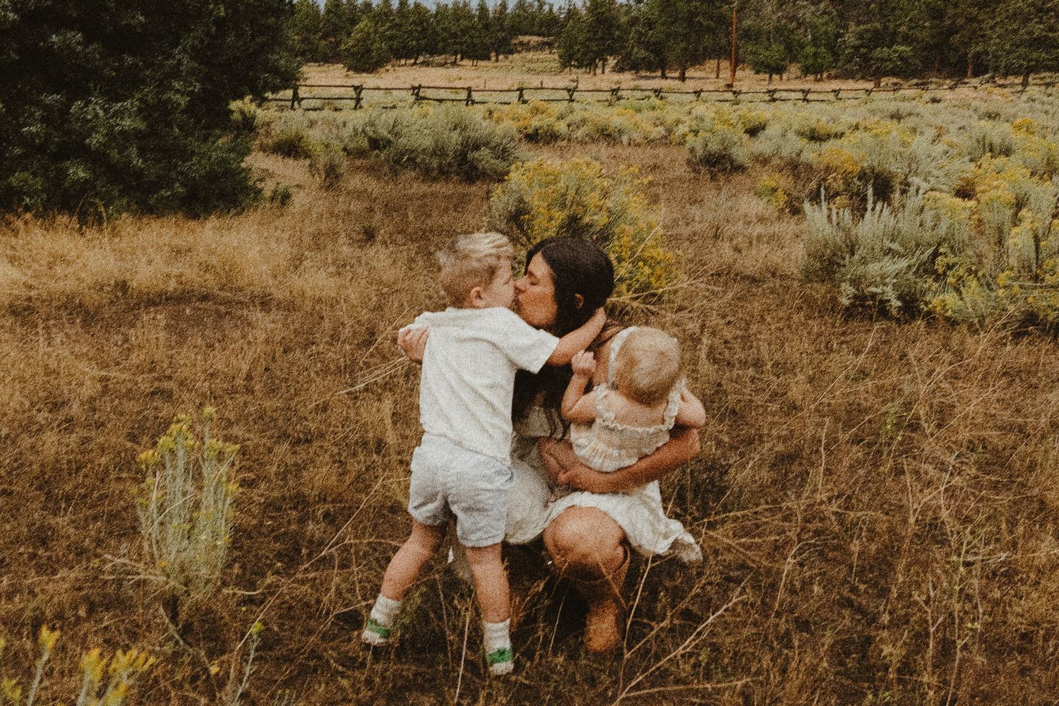 Loving mother crouching in dry grass field hugging two young toddler children.