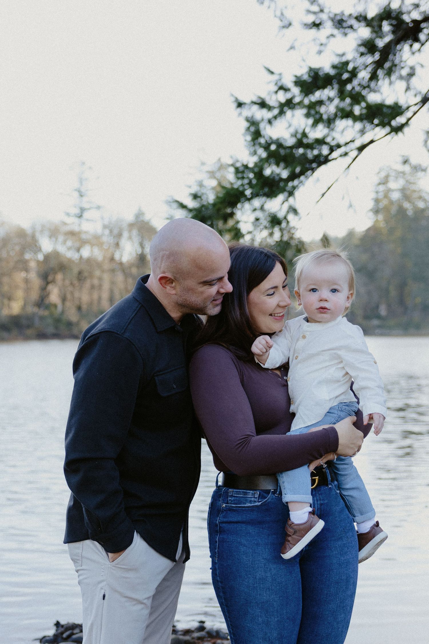 Loving family portrait with parents and baby by a peaceful lake with forest backdrop.