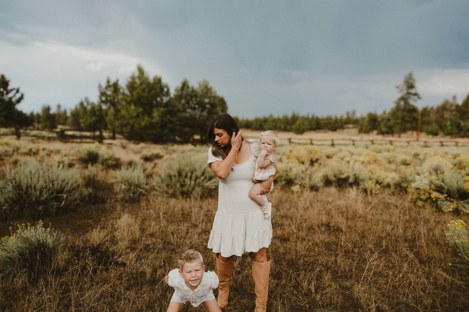 Young mother in white dress lifts baby while toddler plays in golden grass field outdoors.