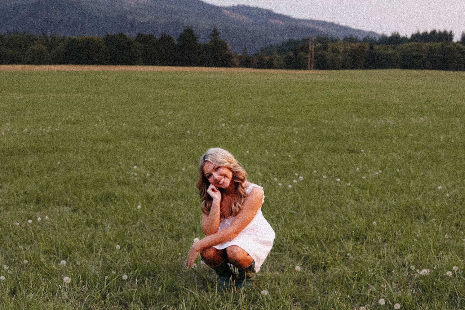 Young blonde girl crouching in green meadow field with mountains and trees in background.