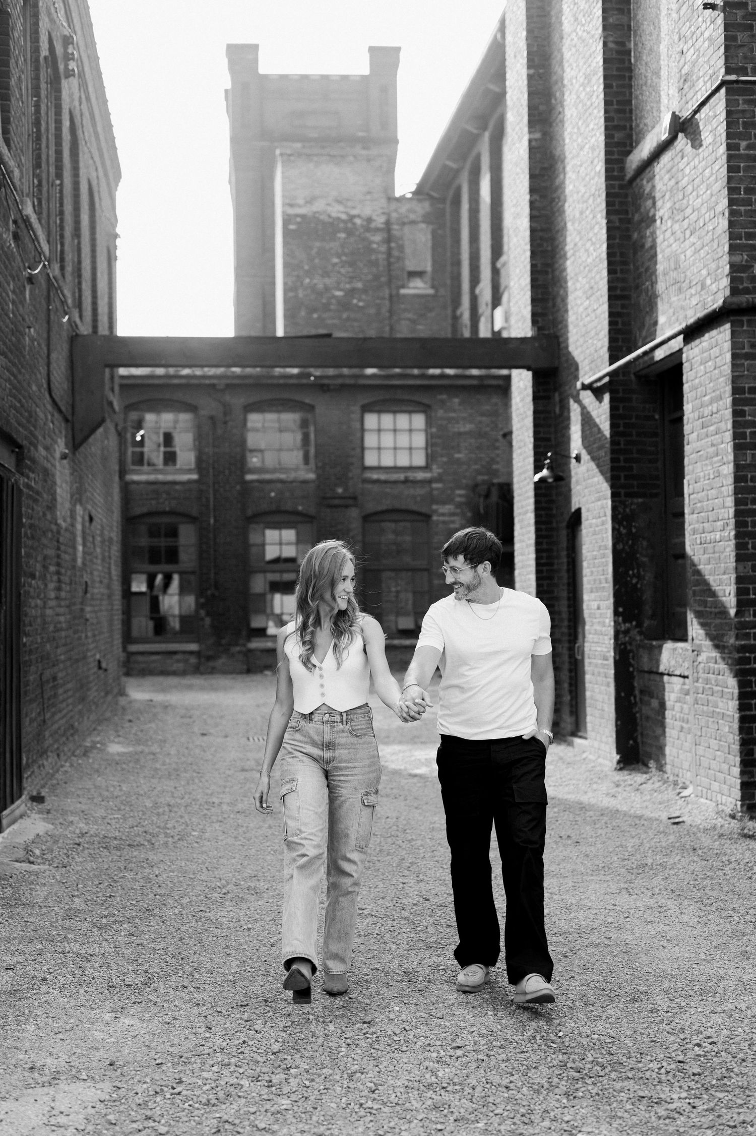 Black and white photo series showing a couple walking hand in hand along a gravel path between industrial buildings.