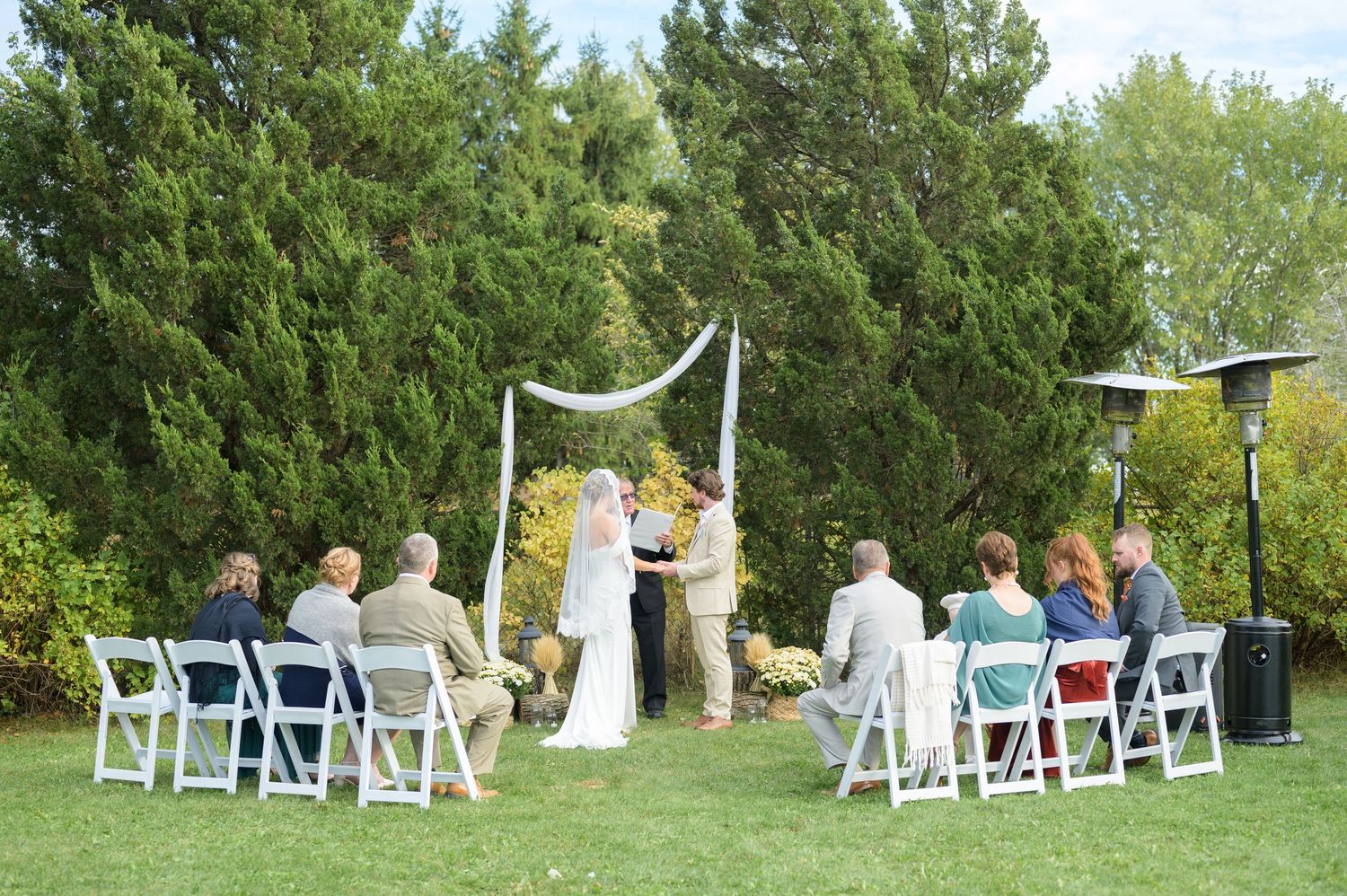White folding chairs arranged for an outdoor wedding ceremony with trees in the background.