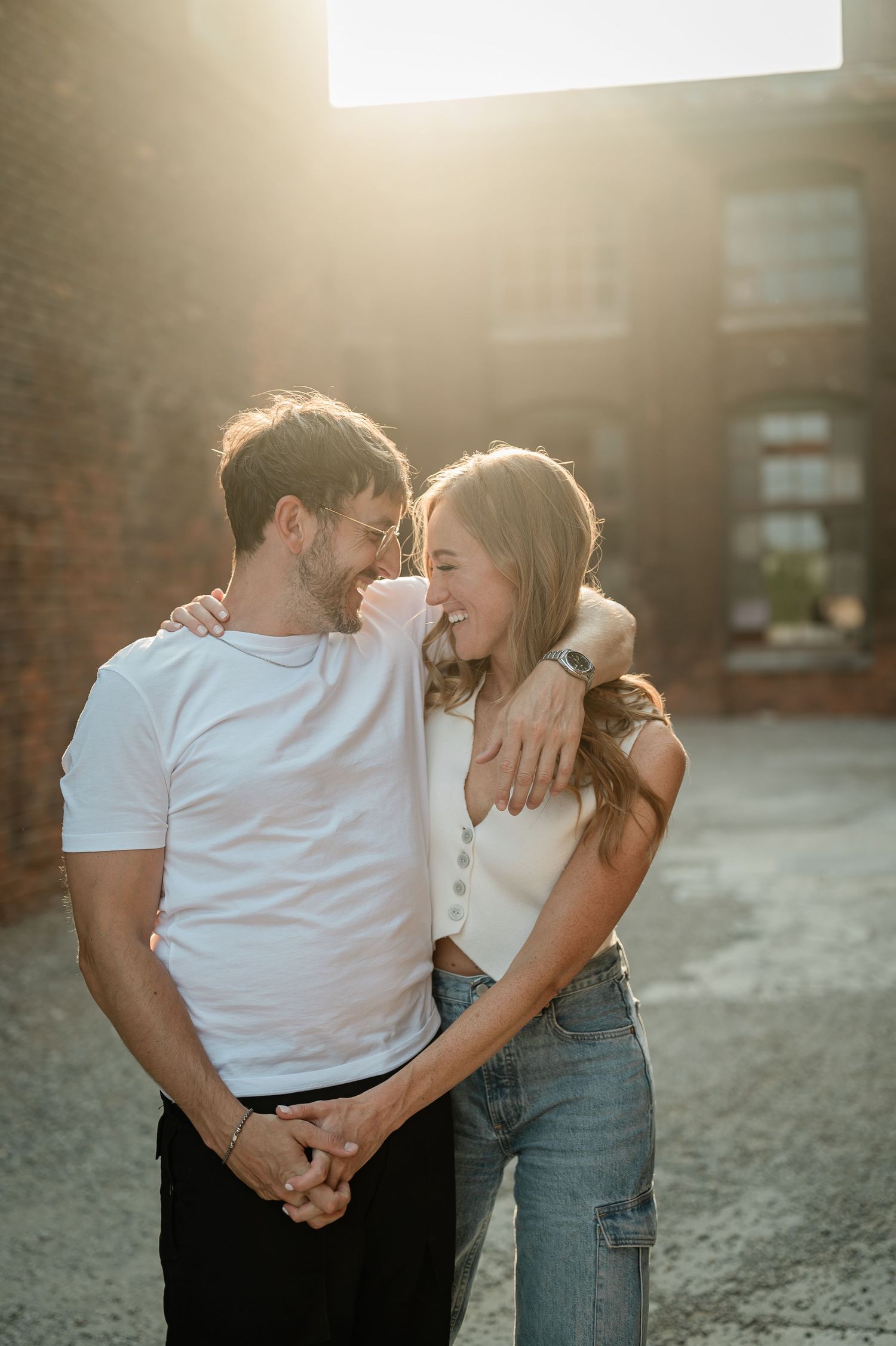 A couple shares intimate moments together against a brick wall backdrop in warm sunlight.