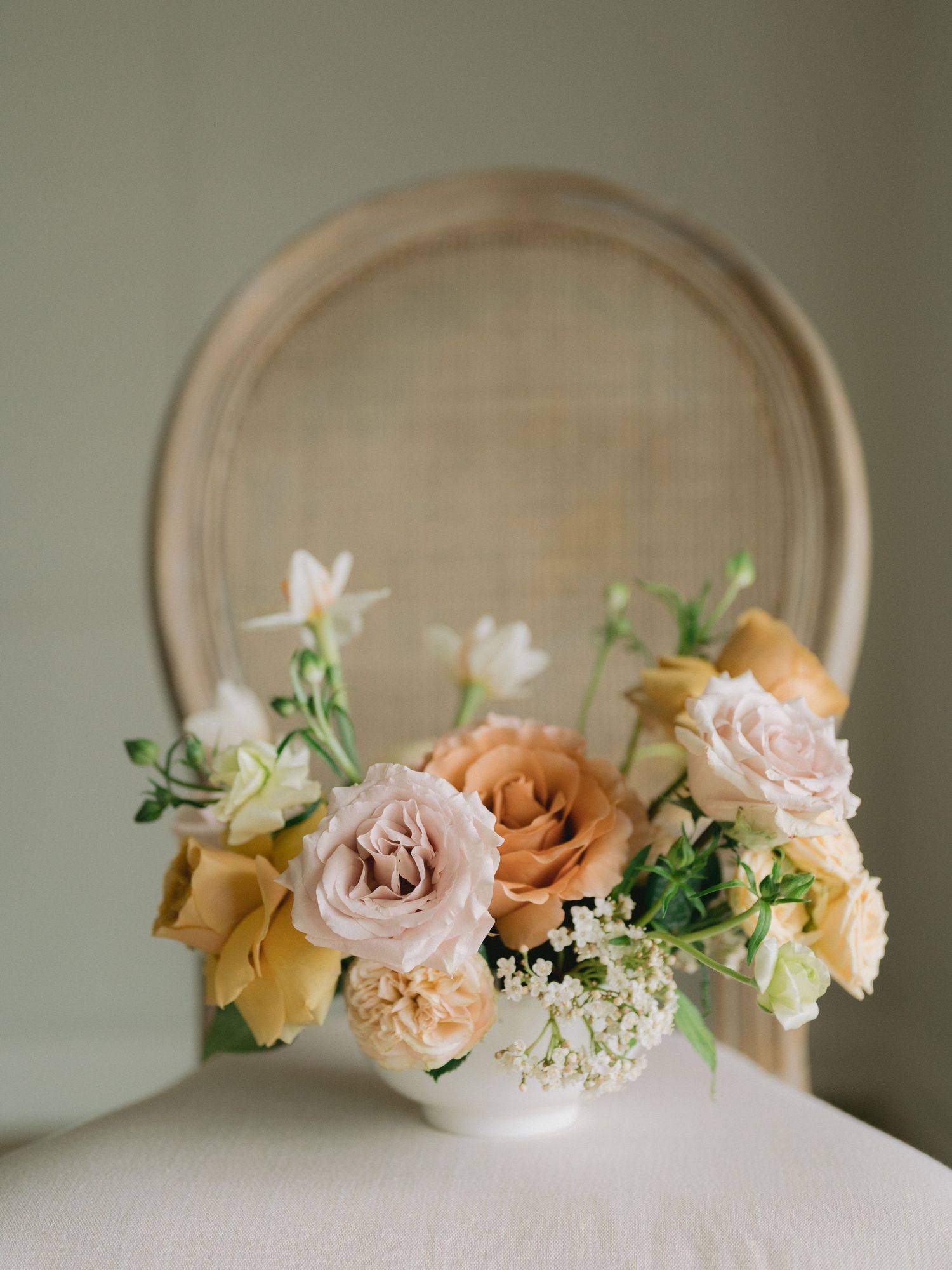 Delicate floral arrangement with peach and blush roses, baby's breath, and white blooms in a white vase against a neutral backdrop.