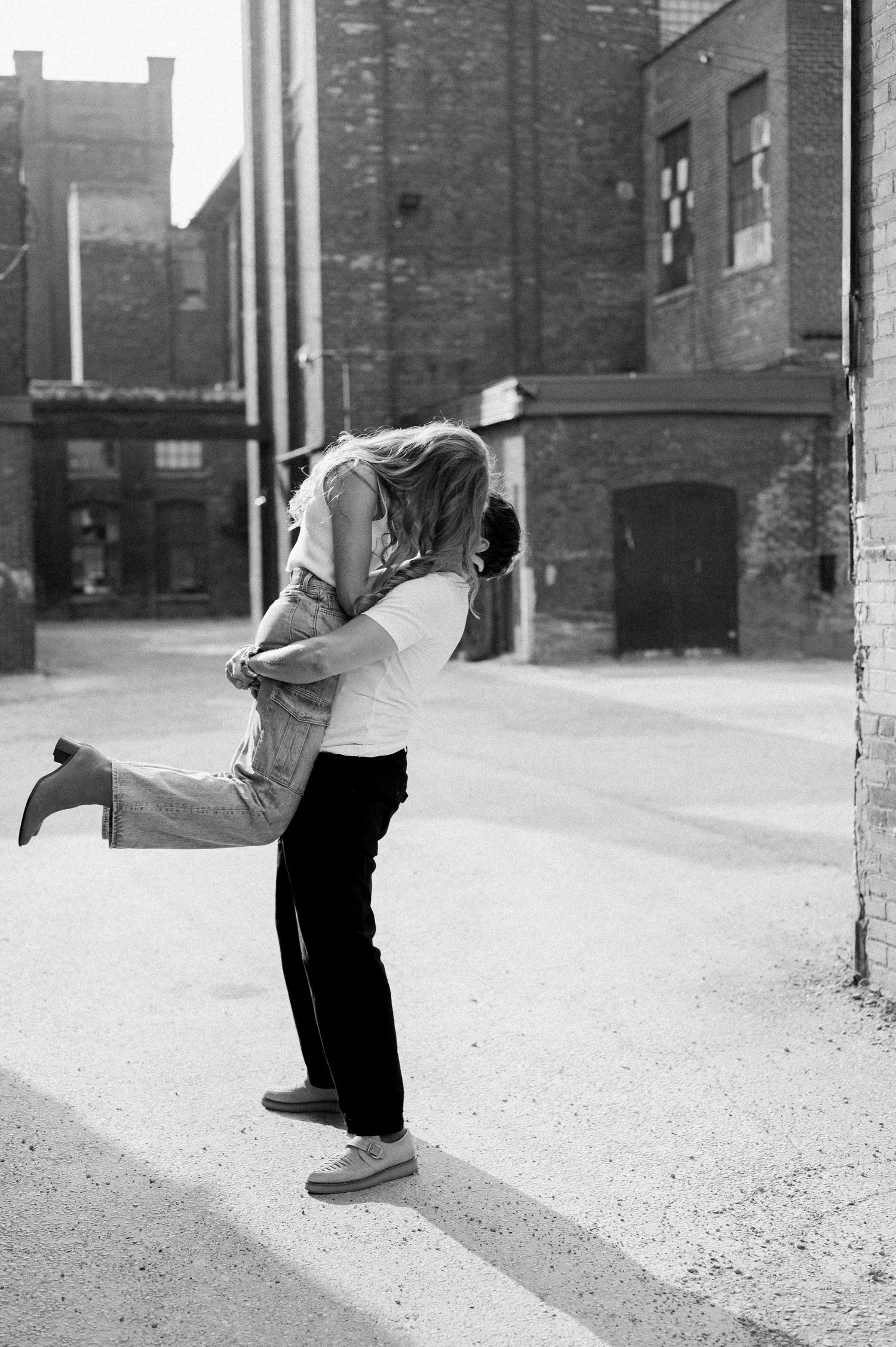A romantic black and white photograph of a couple embracing in an urban alley with brick buildings in the background.