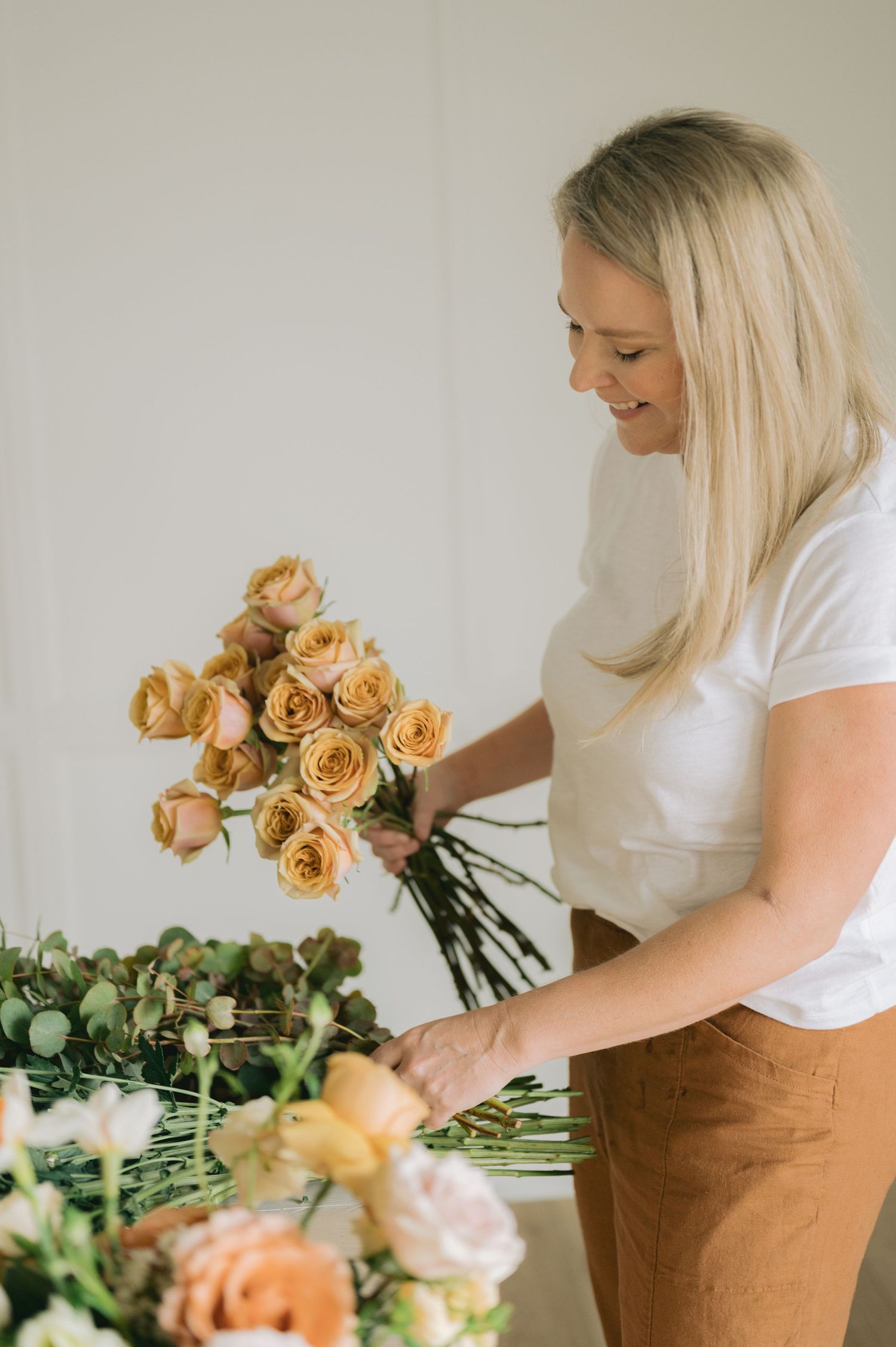 Florist in white shirt and brown pants working with peachy roses and greenery on wooden table.