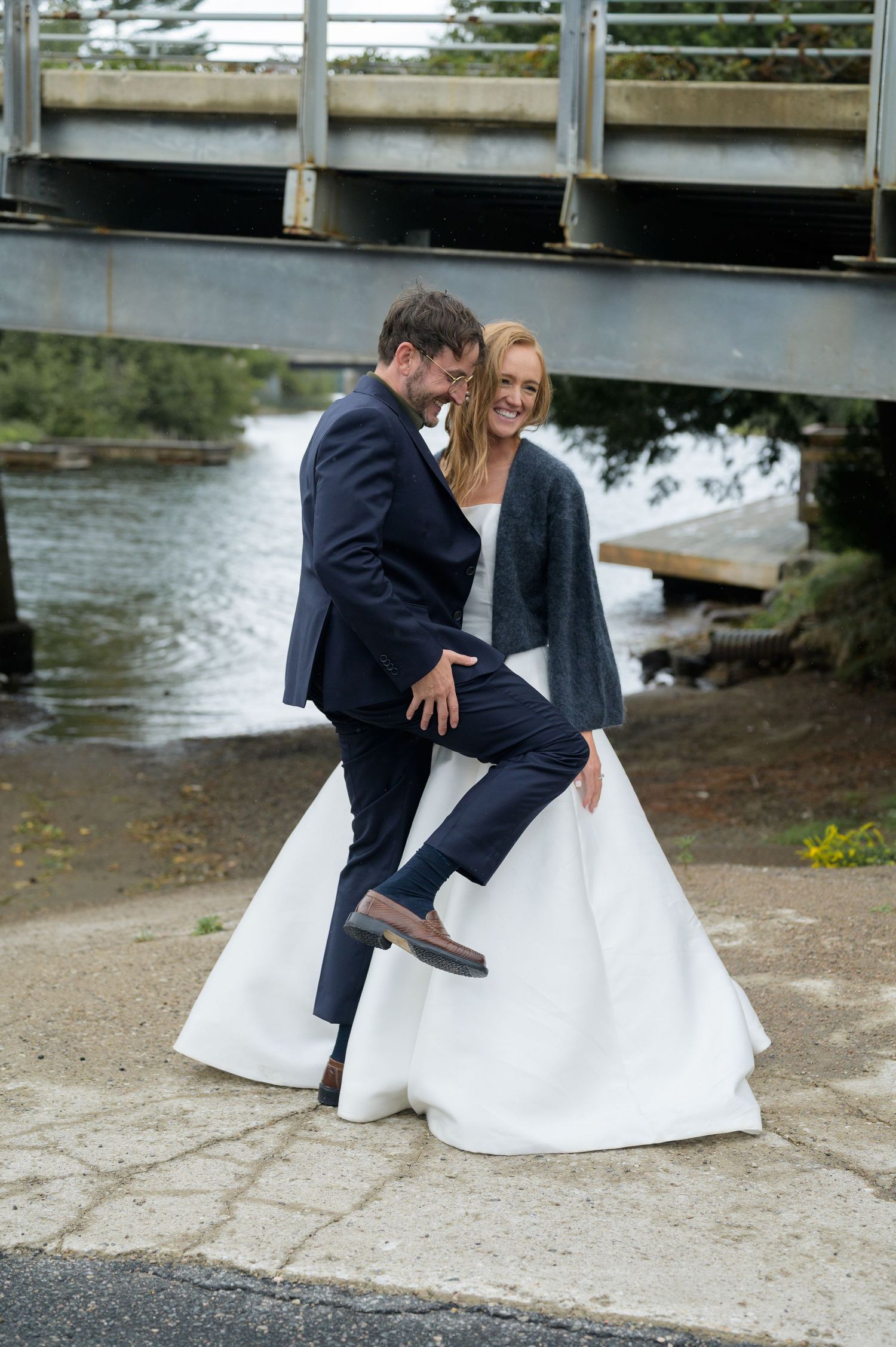 A couple in wedding attire shares a playful moment under a bridge near a waterfall.