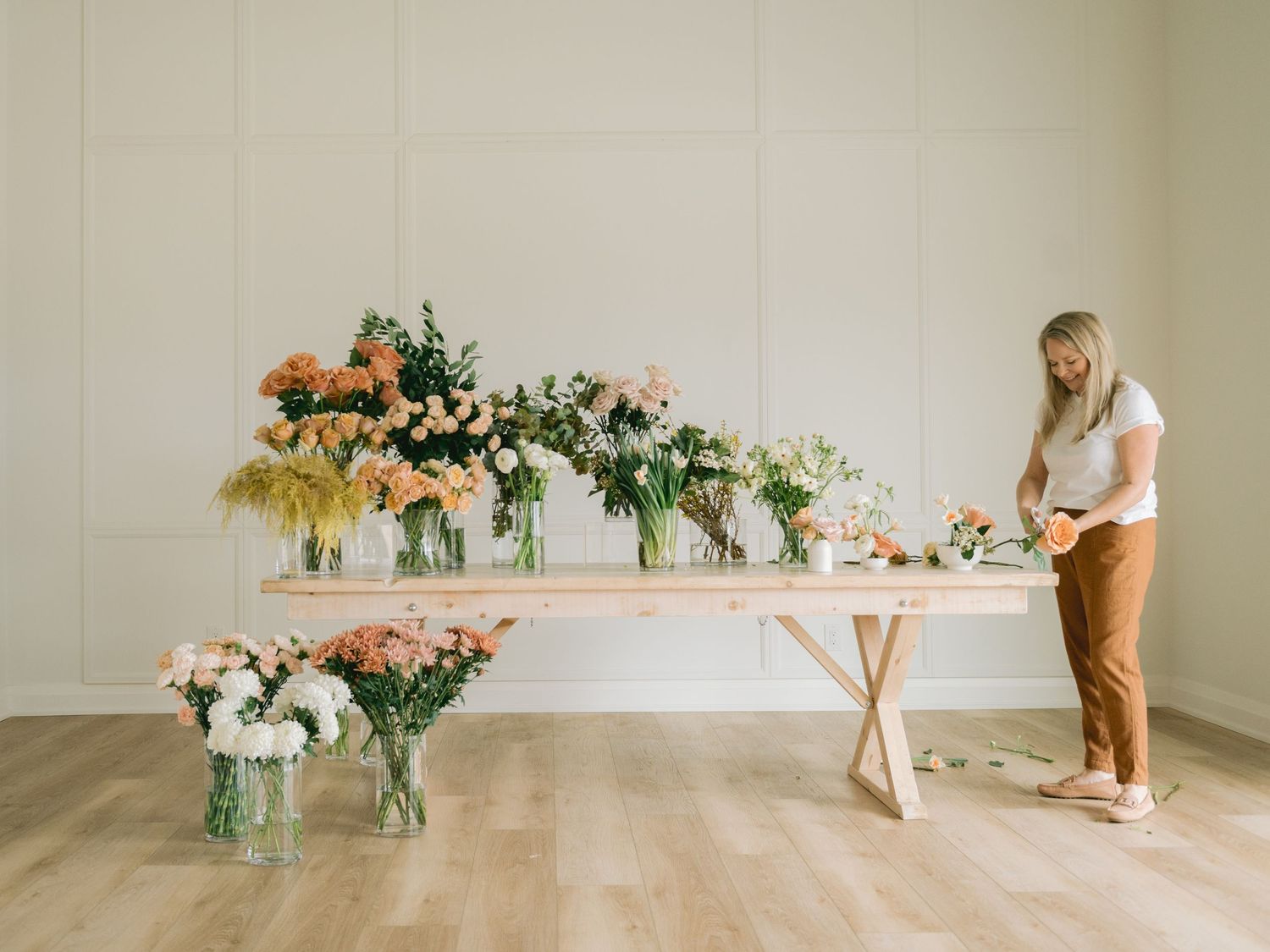 A floral designer arranges fresh flowers on a long wooden table with sunflowers and roses in glass vases.