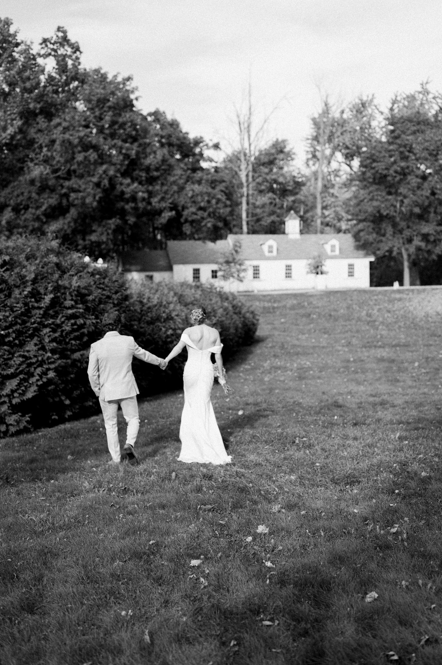 A couple walks hand in hand across a grassy field with white buildings in the background.