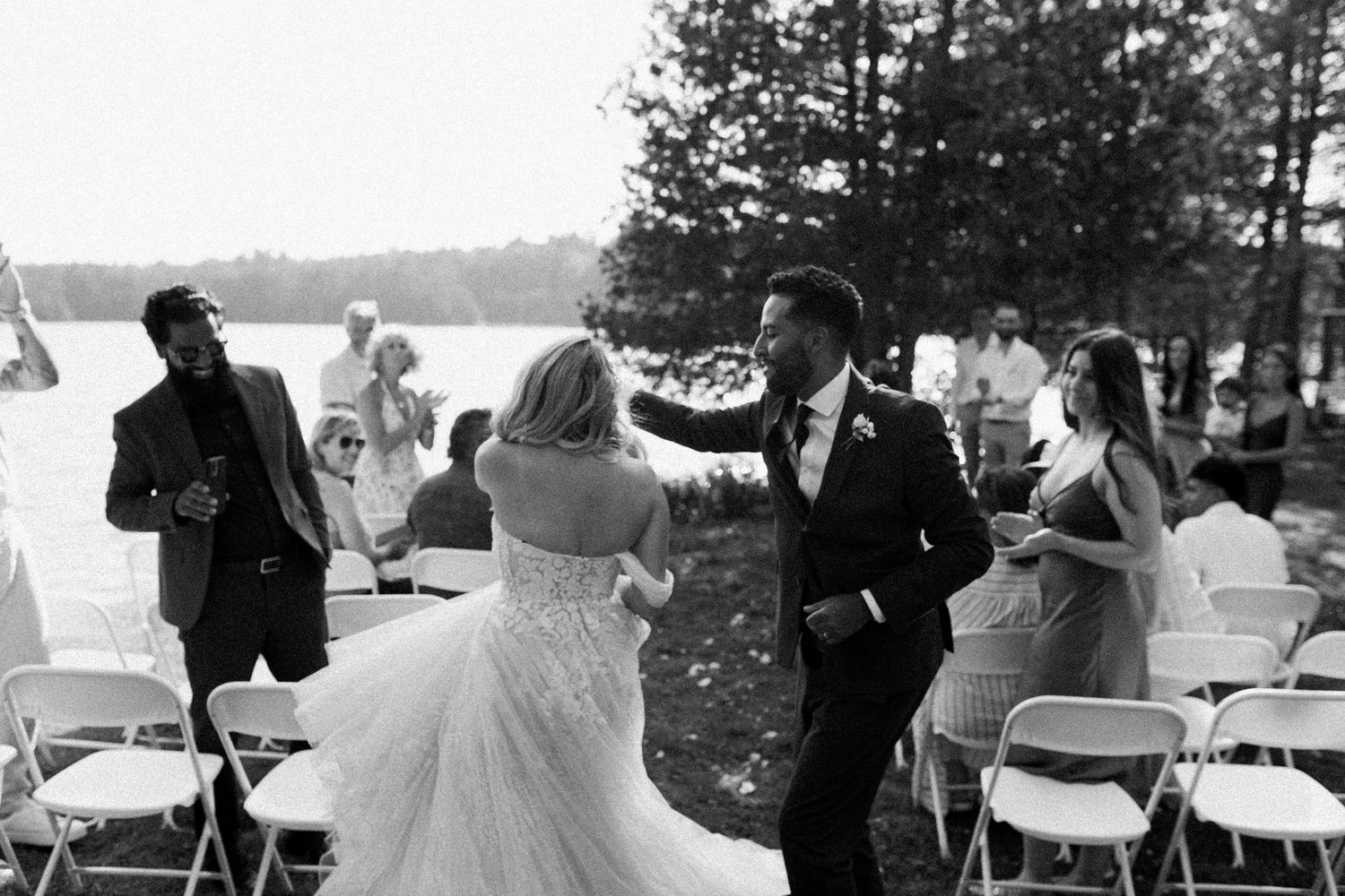 A black and white photo of people dancing and celebrating at an outdoor wedding reception by a lake.