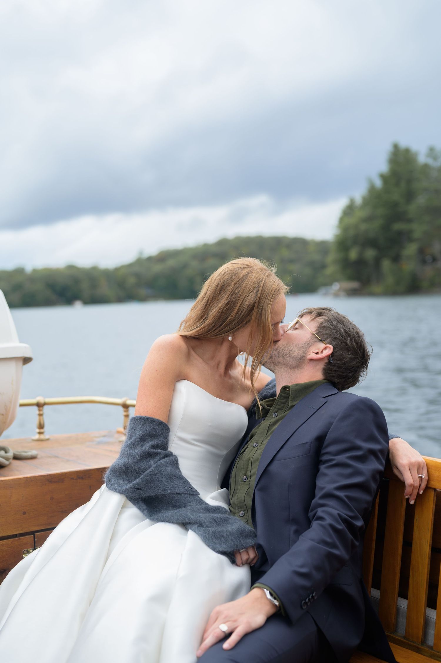 A couple in wedding attire share a romantic kiss on a wooden dock overlooking a serene lake surrounded by trees.