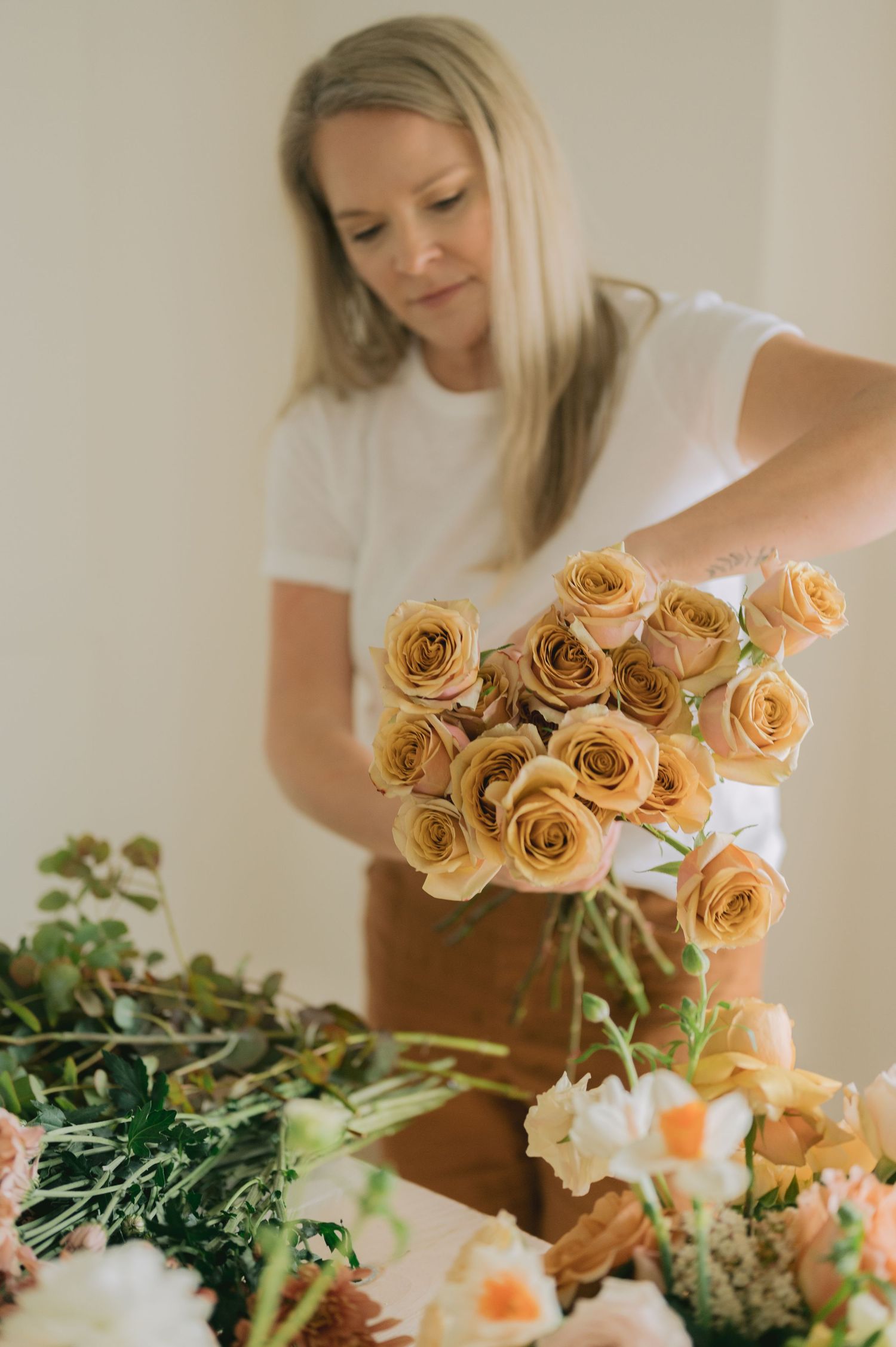 A florist arranges peachy orange roses in a soft, natural light setting with greenery on a wooden surface.