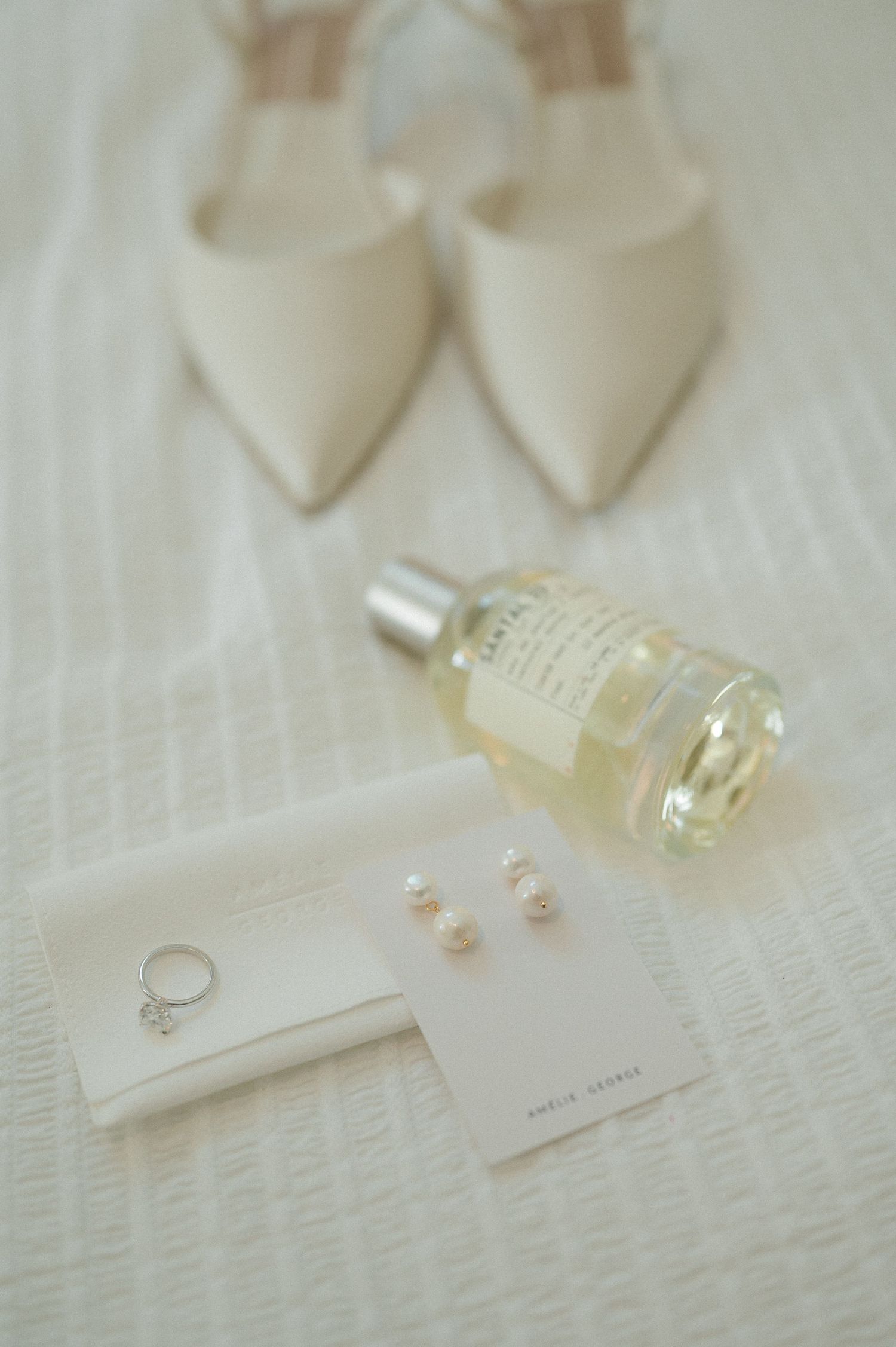 Elegant wedding details featuring perfume bottles, pearls, and delicate lace on a soft white textured surface.