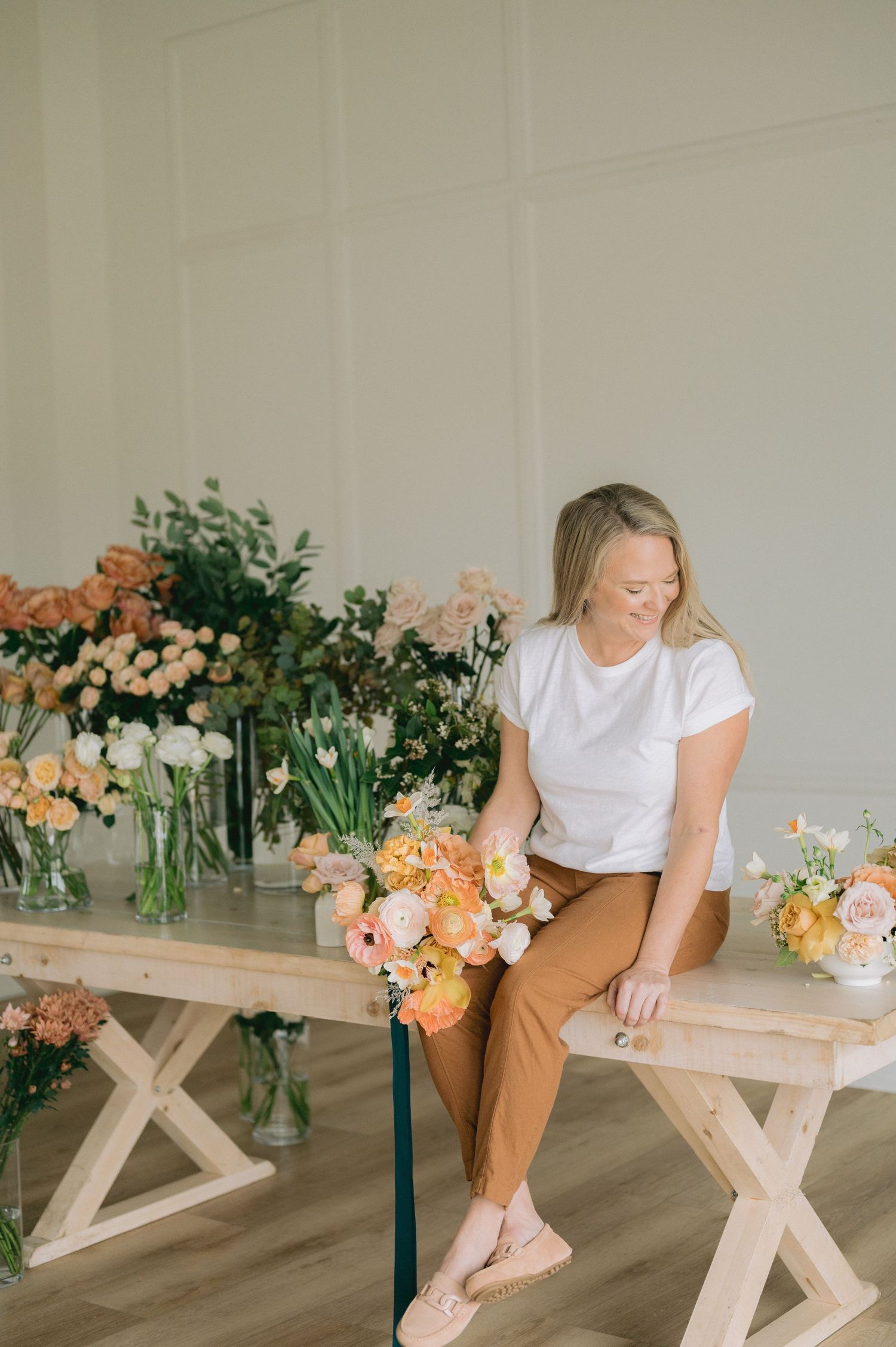 Person arranging peach and cream colored flowers on a rustic wooden bench in minimalist white room.