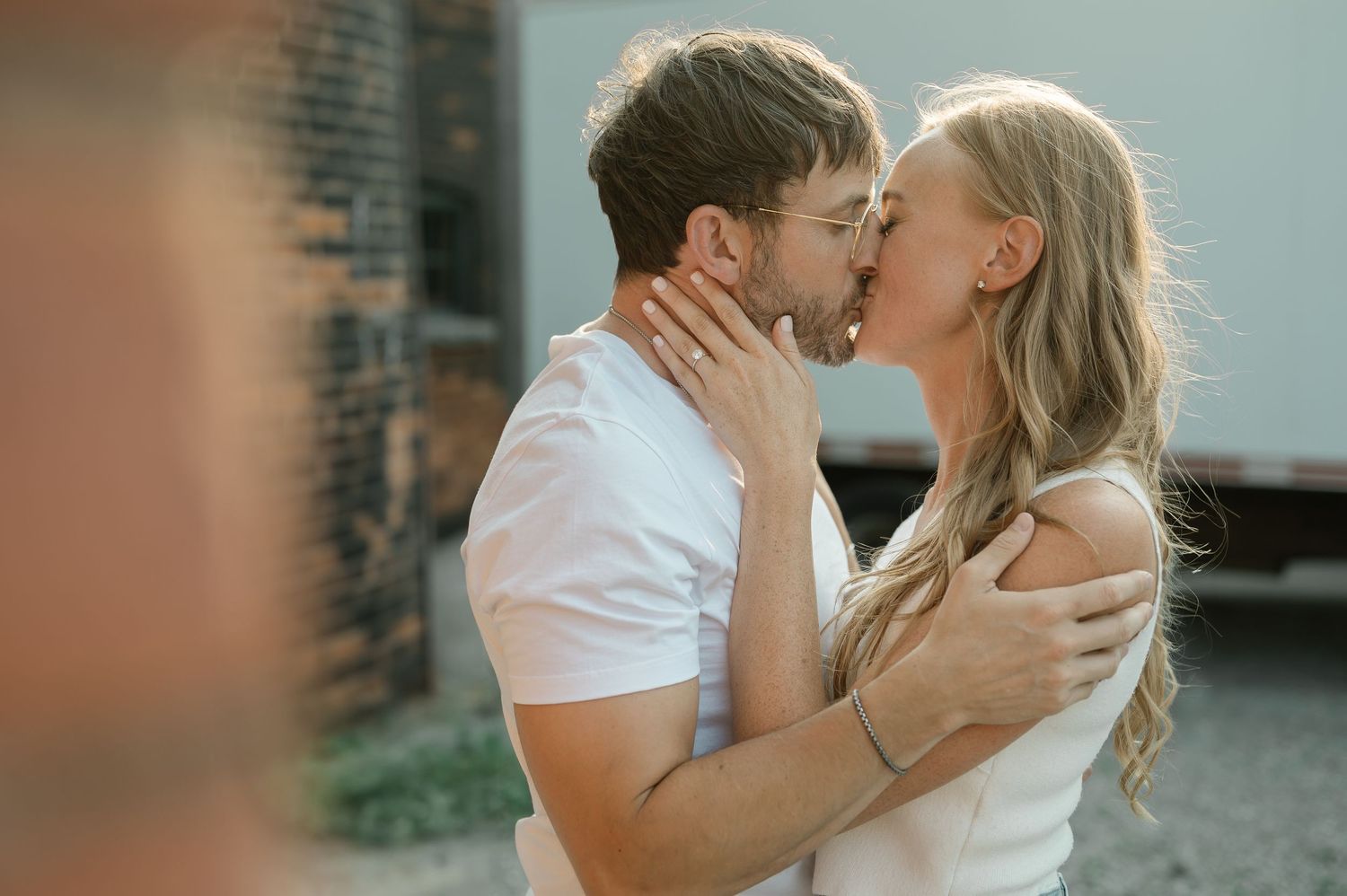 A romantic couple in white outfits share a tender kiss outdoors against a brick wall background.