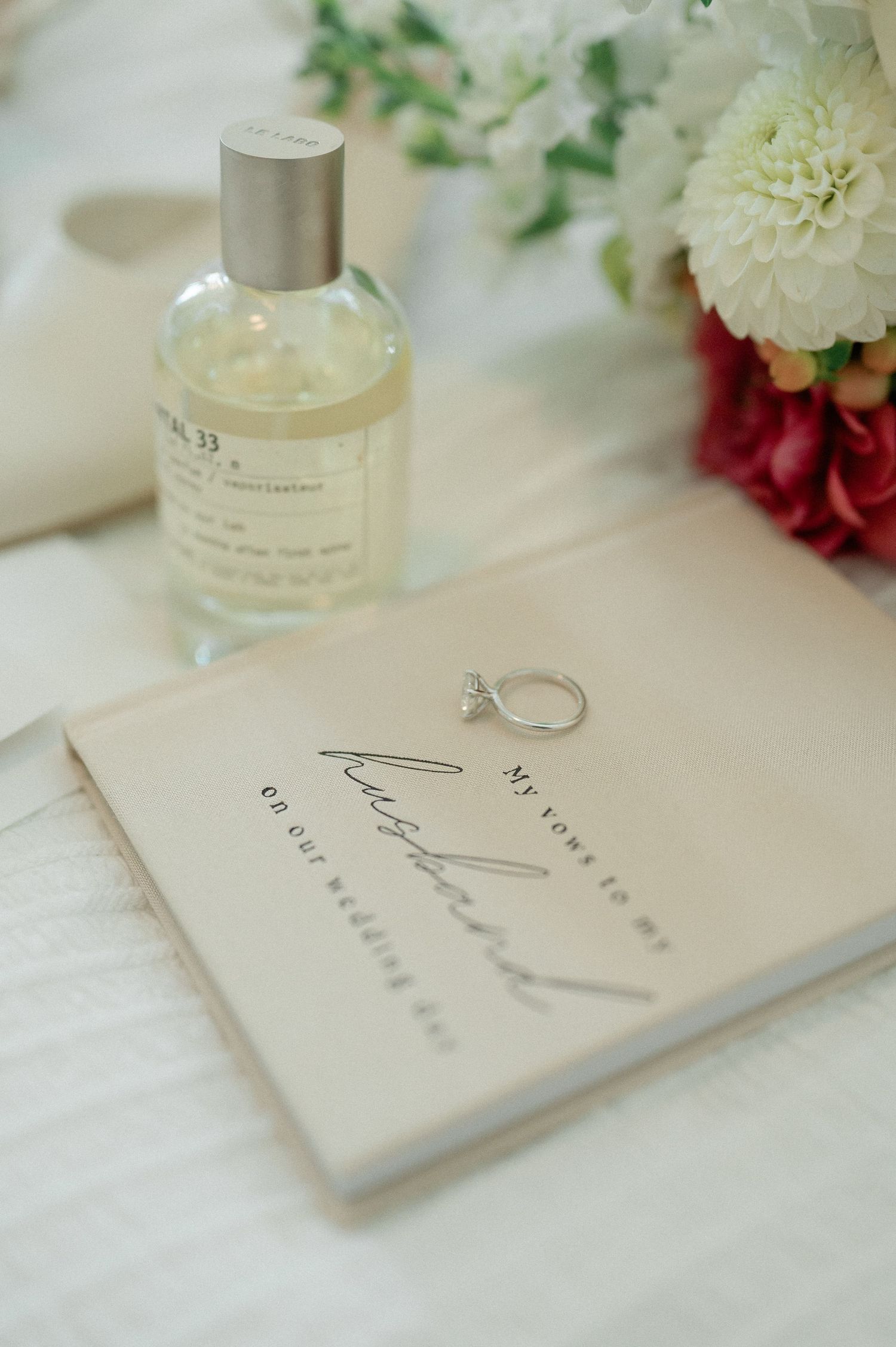 A wedding ring sits beside a perfume bottle and book on a white surface with pink and white flowers in the background.
