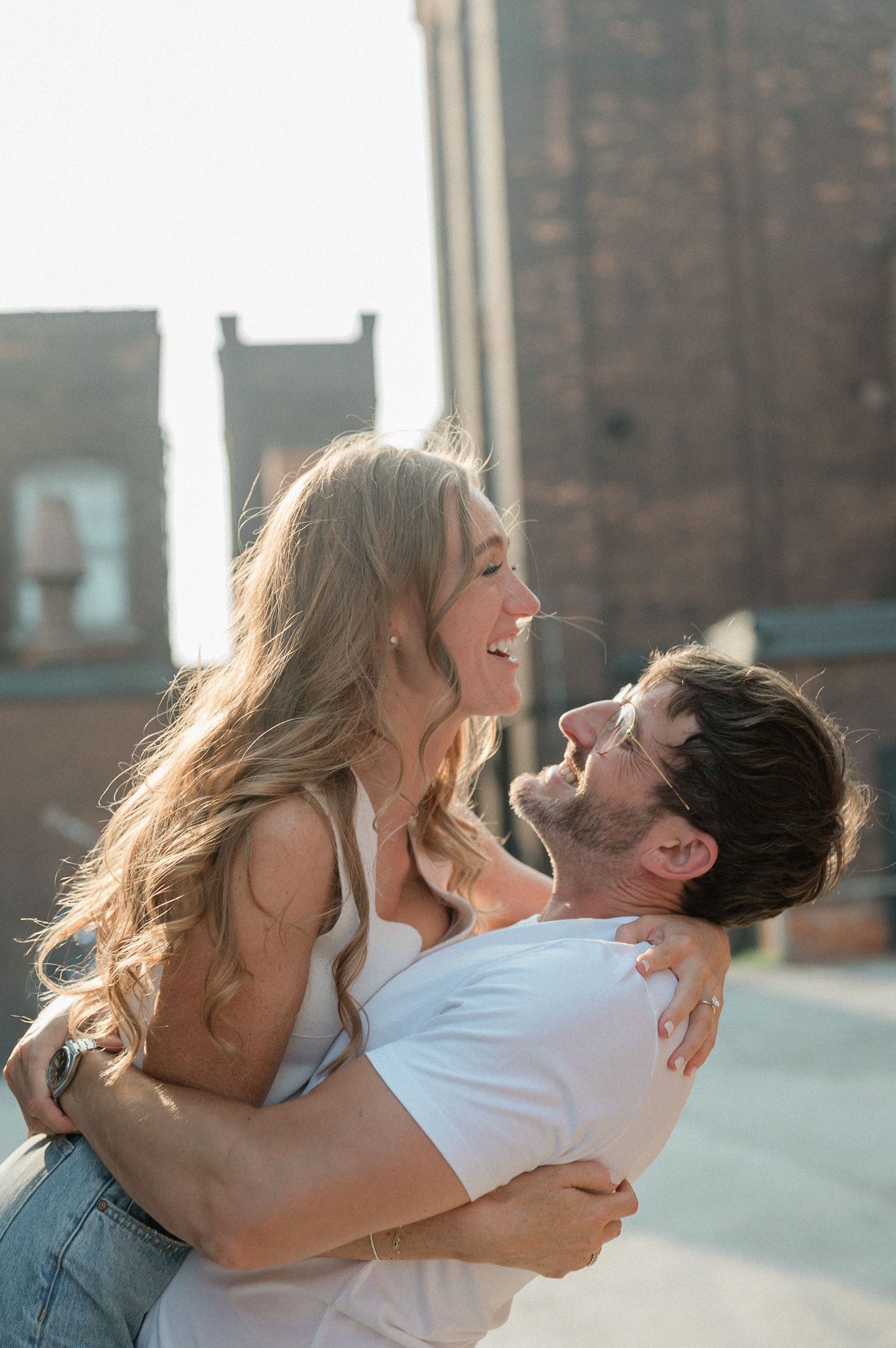 A joyful couple embraces and laughs together on a rooftop with urban buildings in the background.