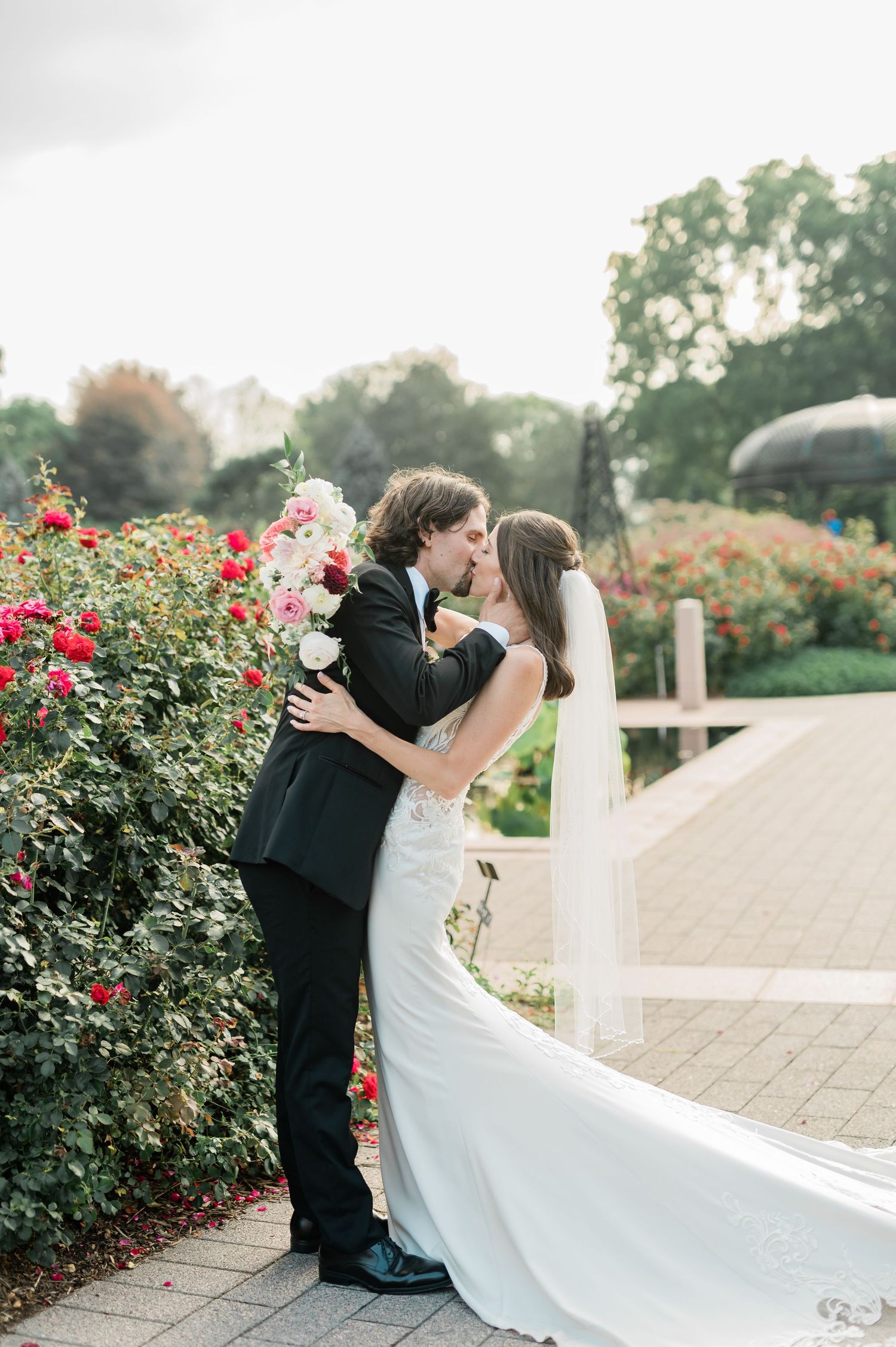 A romantic wedding couple shares a kiss in a garden surrounded by blooming red roses on a stone pathway.
