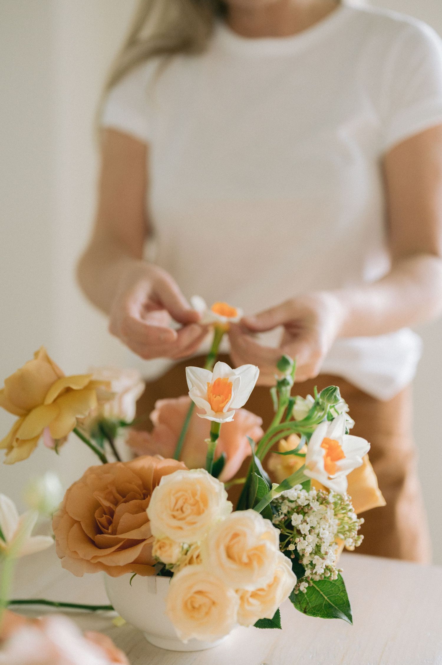 A delicate floral arrangement of peach roses and white daffodils in a white vase being arranged by hands in white.