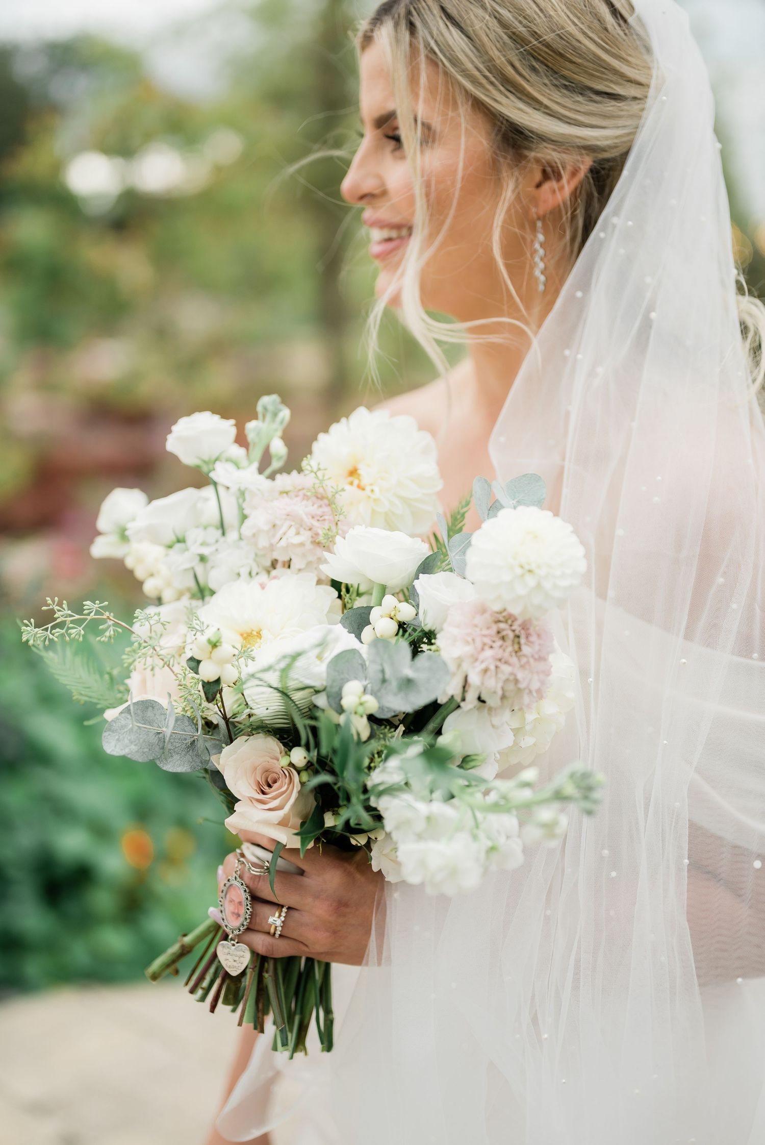 A bride in a white veil holds a romantic bouquet of blush roses, white dahlias and delicate greenery.
