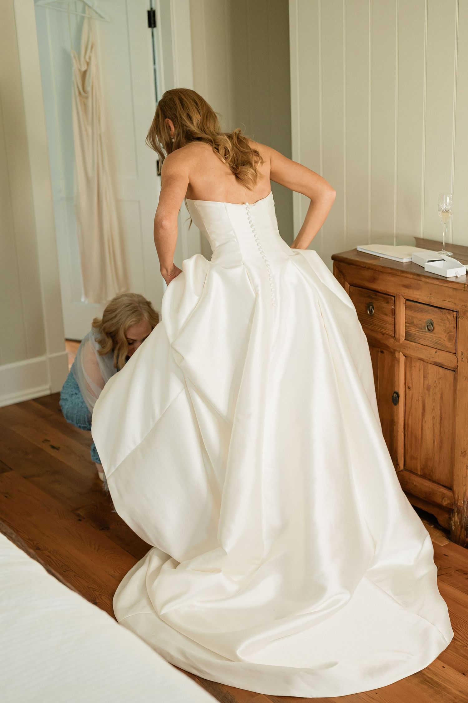 A bride in a flowing white wedding gown twirls gracefully on a wooden floor beside rustic furniture.