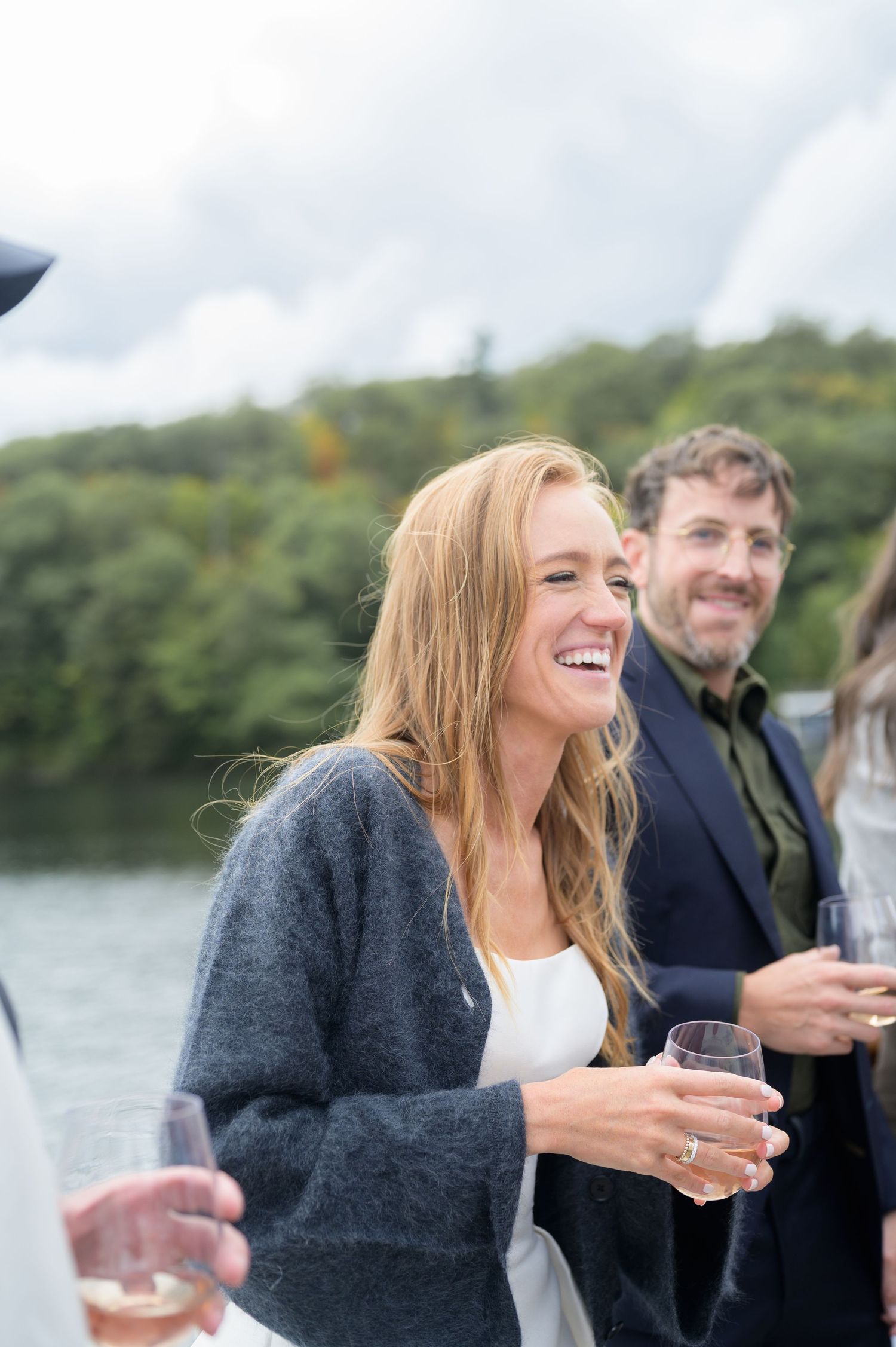 A group of friends enjoying drinks and laughter by a scenic lake on an overcast day.