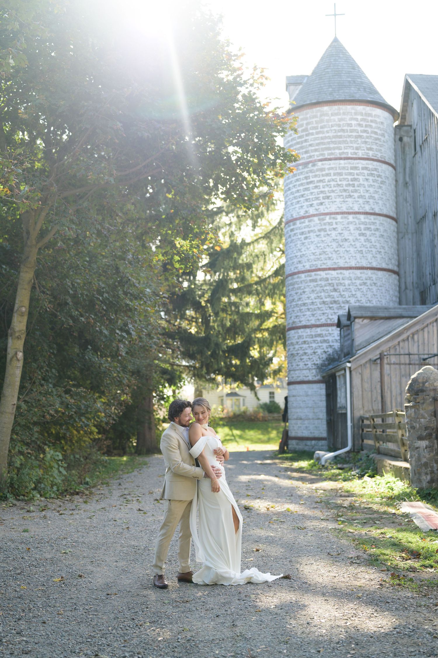 Newlyweds embrace near a historic stone tower on a sunlit forest path.