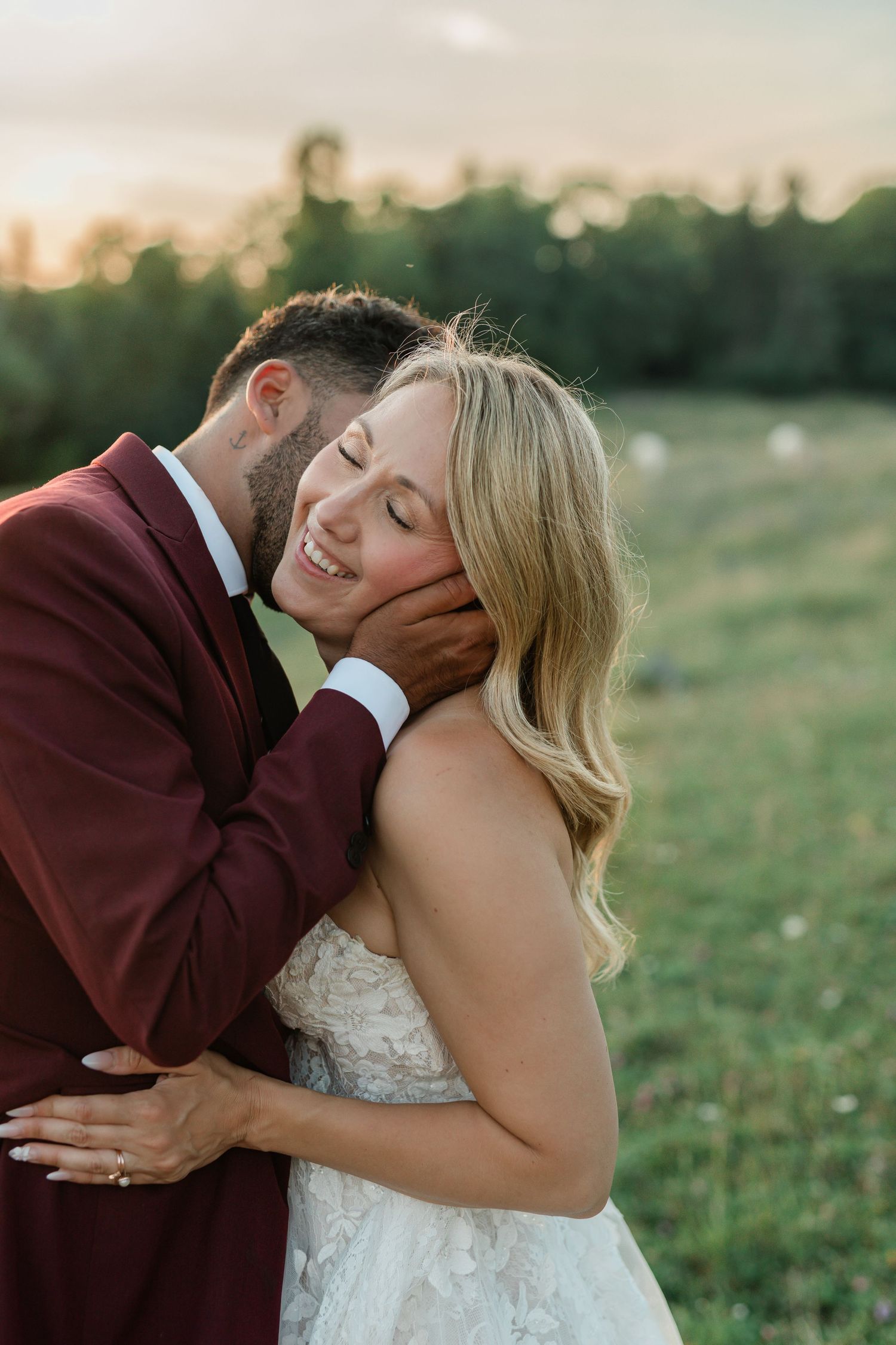 Romantic couple sharing intimate kisses in an outdoor field at sunset wearing burgundy and white wedding attire.