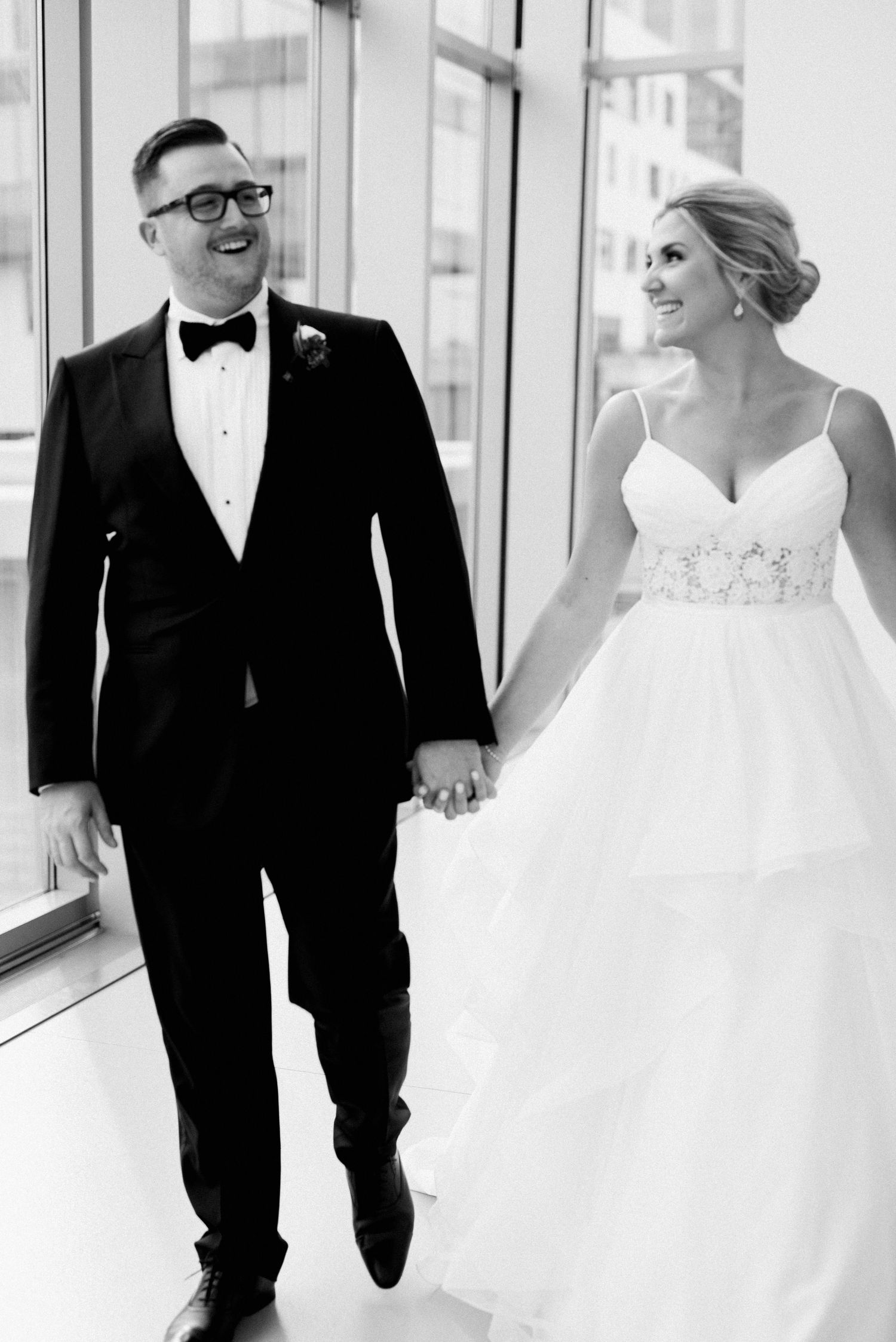 Black and white wedding photo showing couple walking hand in hand down a bright hallway in formal attire.
