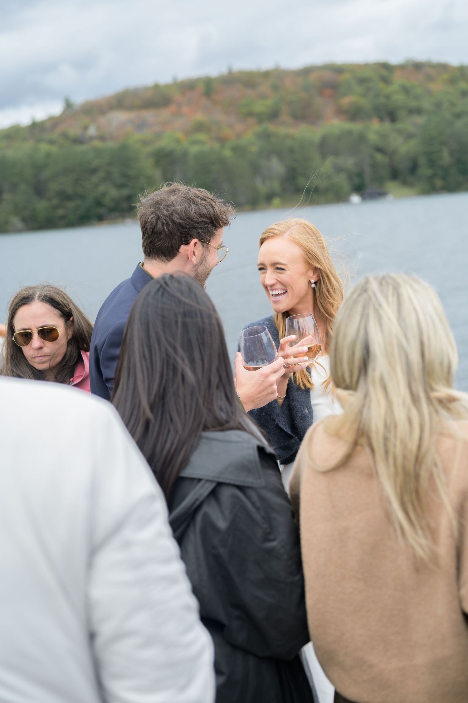 Friends gathered outdoors sharing wine and conversation against a backdrop of autumn foliage and water.