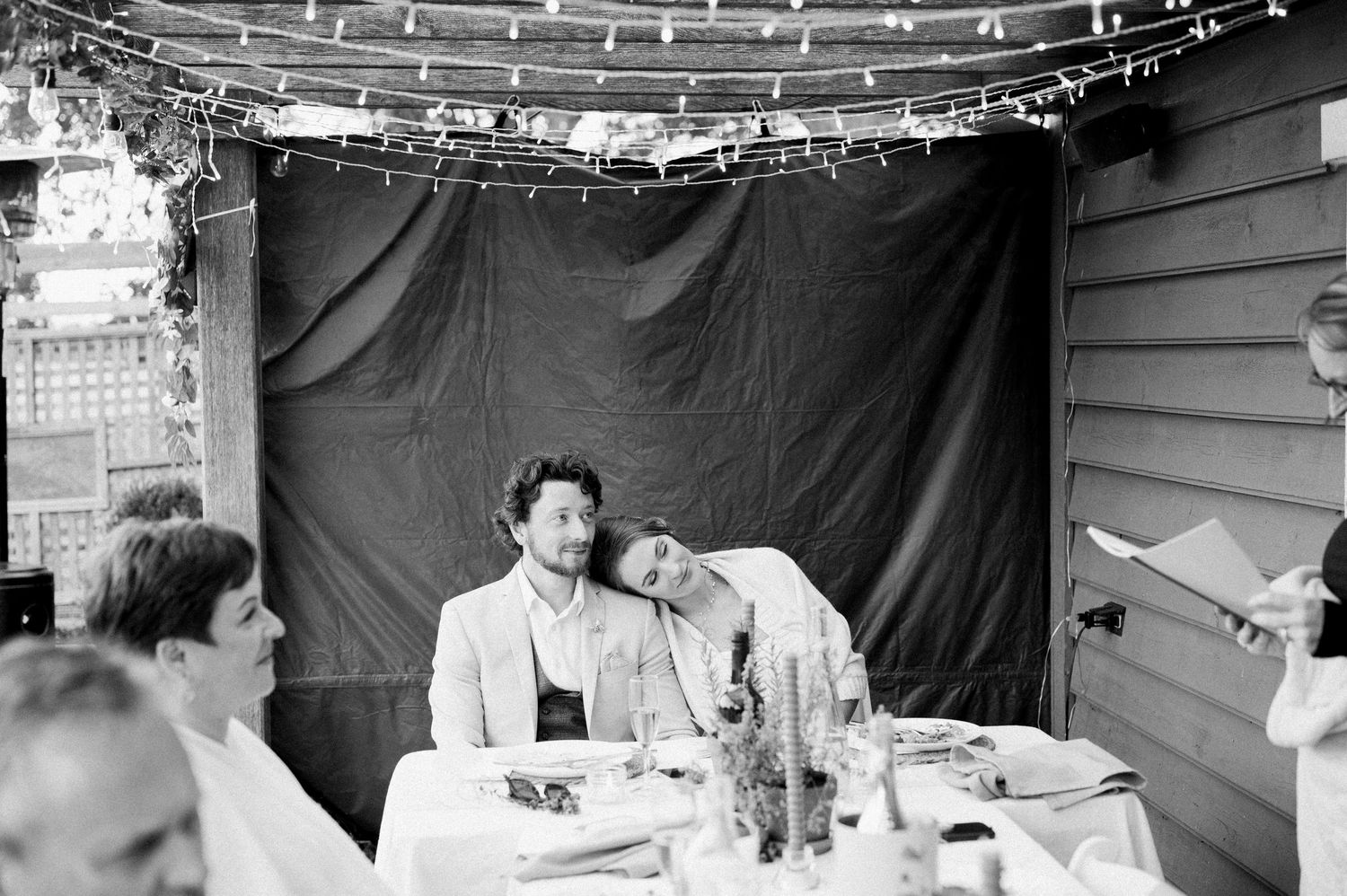 A black and white photo of people seated at a dinner table with string lights and a dark curtain backdrop.